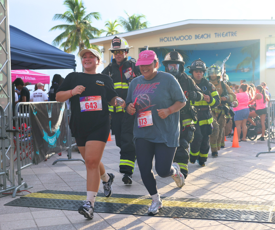 Two smiling women cross the finish area side by side, with a line of firefighters in gear behind them.