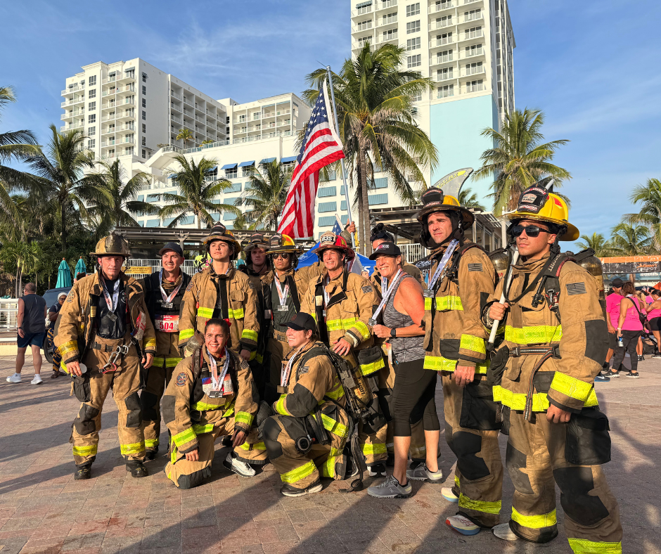 Firefighters in tan gear with medals pose with a few other people and a U.S. flag in front of beachfront hotels.