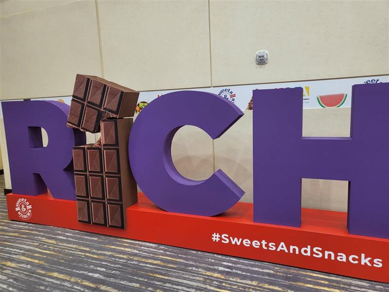 Giant purple letters RICH with a chocolate bar bitten out of the I, above the hashtag SweetsAndSnacks.