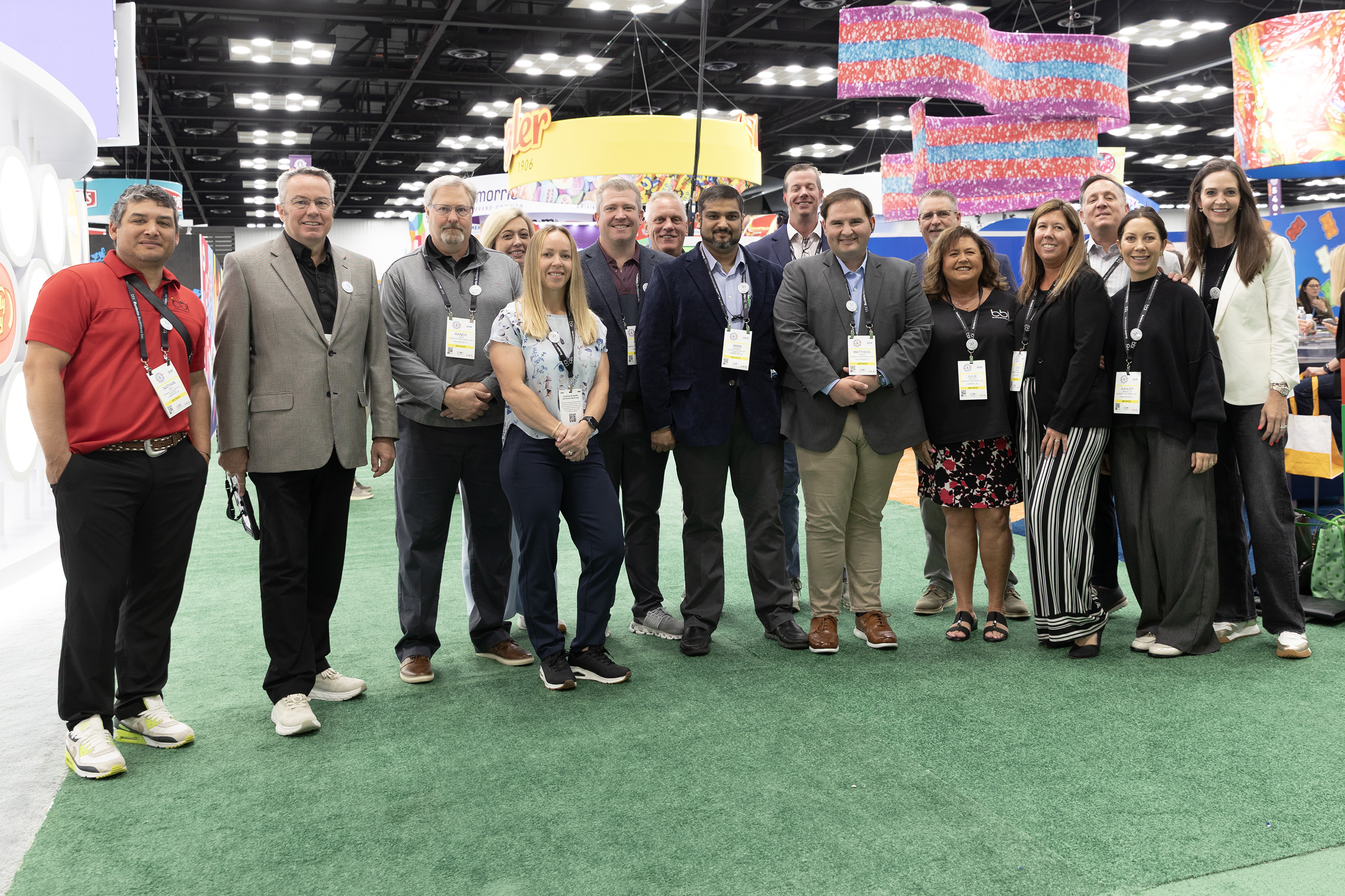 Large group of adults with conference badges poses on green turf under hanging candy-colored signs in an expo hall.