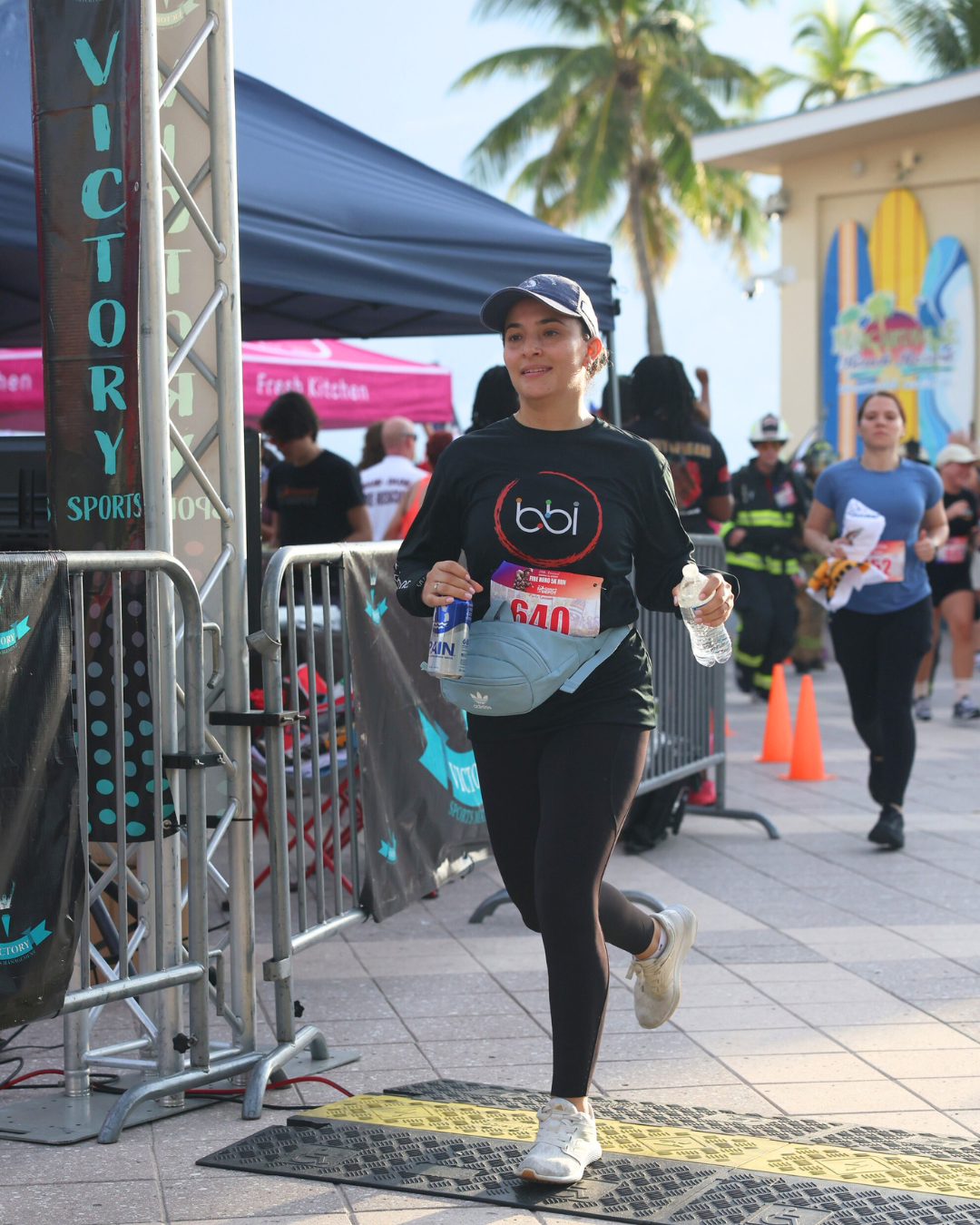 Woman in a bbi shirt runs through the finish area, past barricades and a blue tent, holding a water bottle.