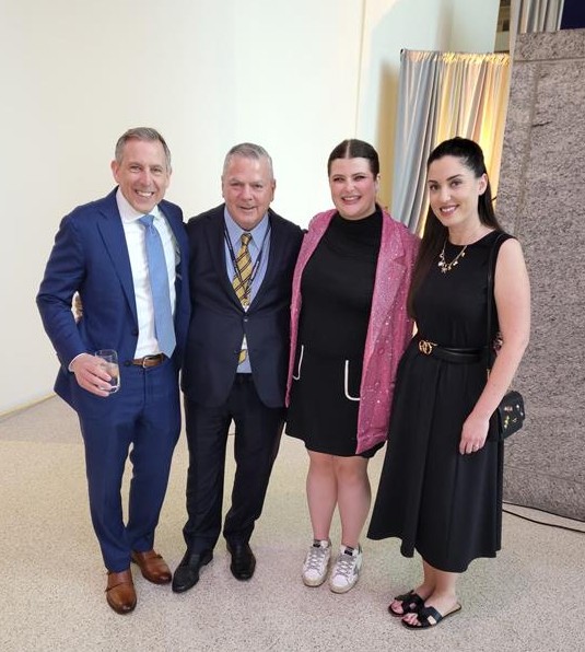 Two men and two women pose together indoors, the man second from left in a striped tie and a woman in a pink sparkly blazer.