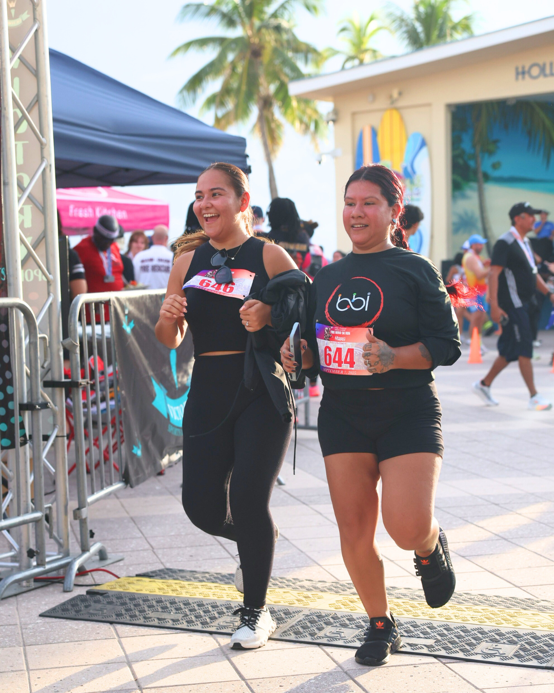 Two laughing women, one in a bbi shirt, jog together past barricades at the finish line.