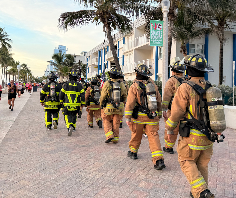 Firefighters in black and tan gear with air tanks walk away from the camera along a palm-lined brick boardwalk.