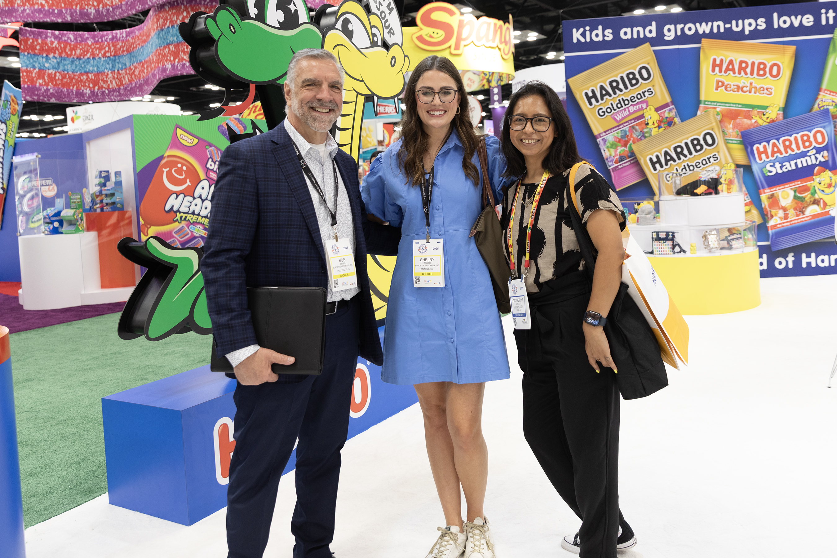 A man and two women with badges pose before a Haribo display and green snake graphics at an expo booth.