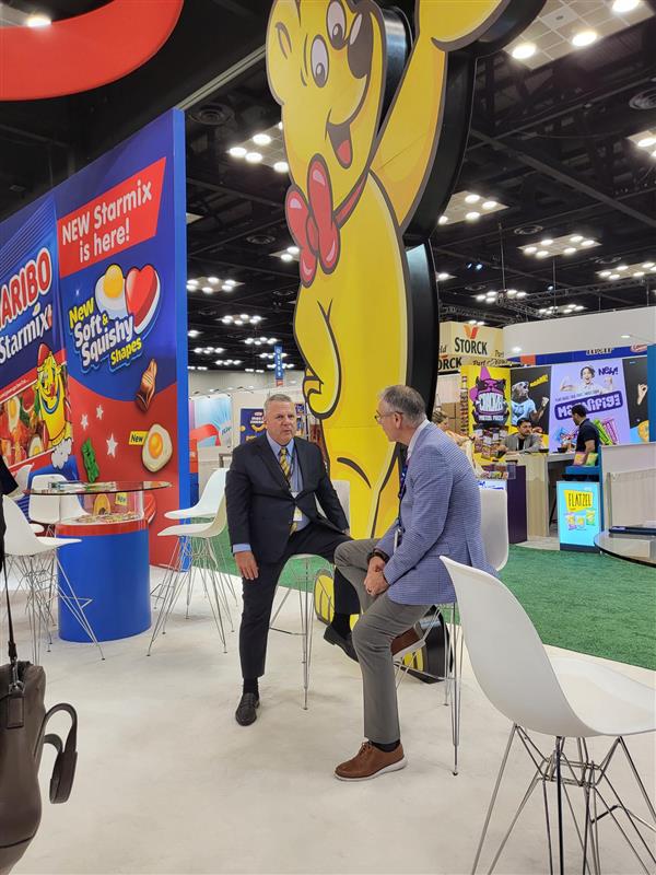 Two men talk seated on white stools in a Haribo booth under a tall yellow gummy bear figure and a New Starmix sign.
