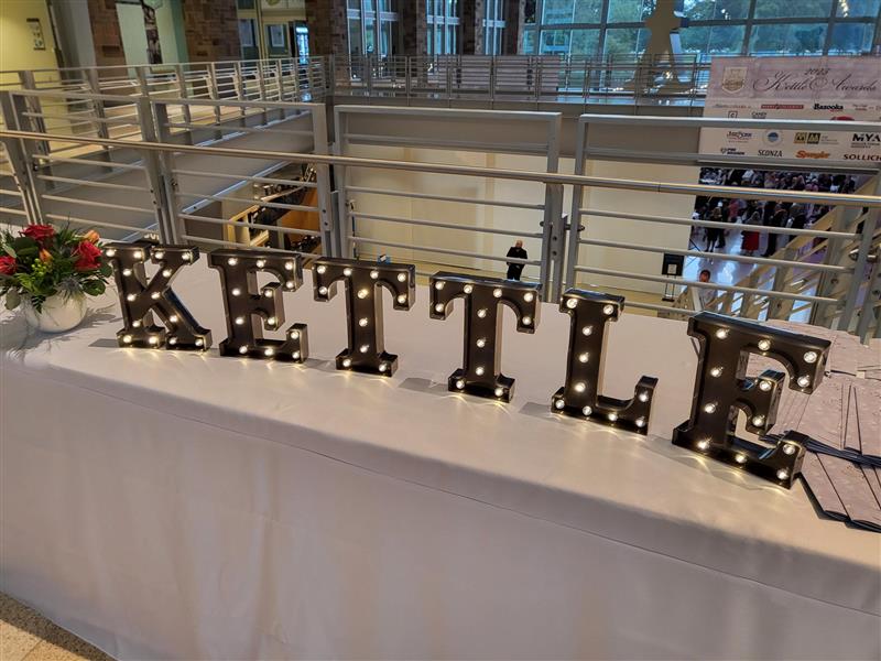 Lit marquee letters spelling KETTLE on a white-draped table inside a conference building atrium.