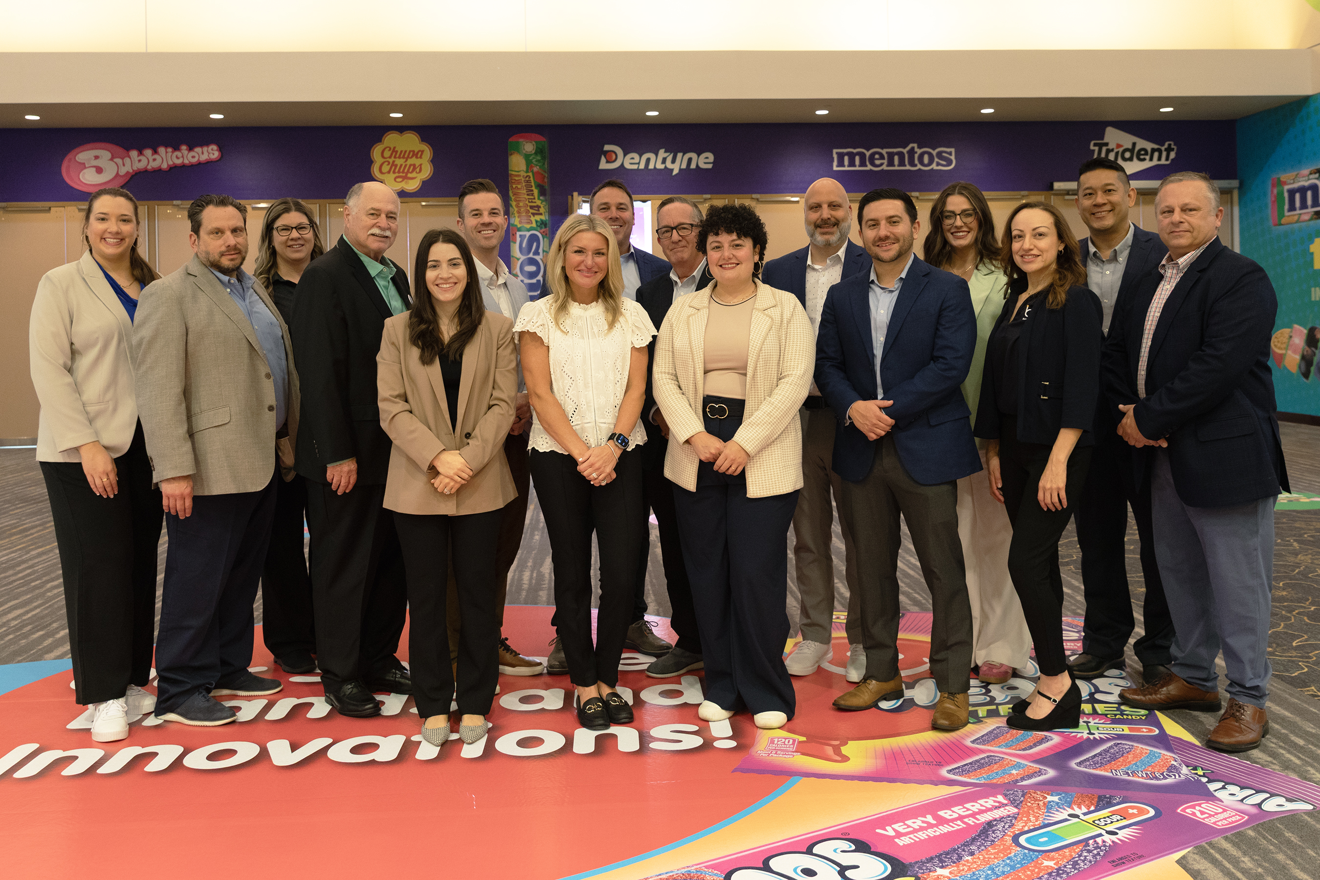 Group of adults in business attire pose on a floor graphic reading Innovations, below Bubblicious, Dentyne and Mentos signs.