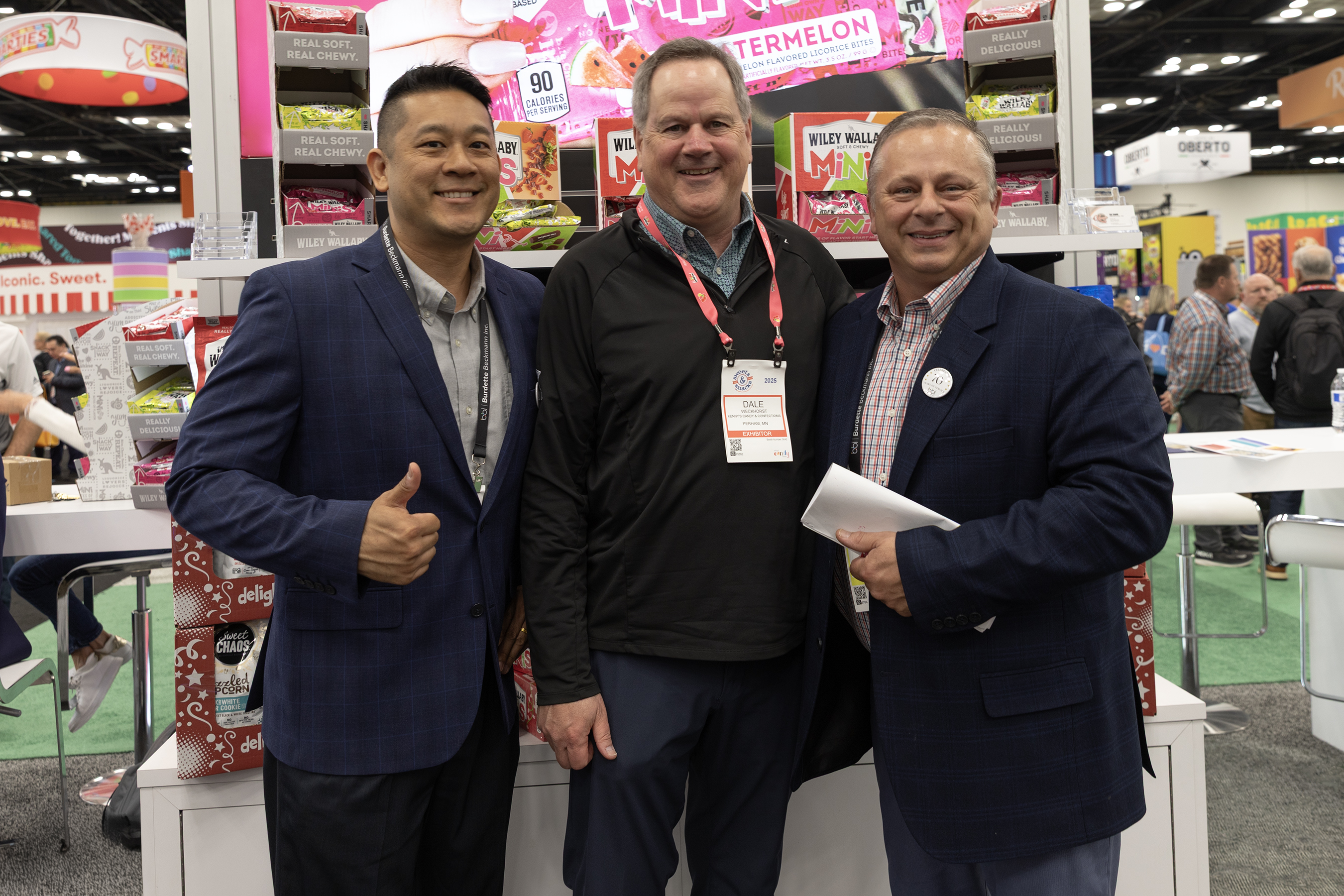 Three men pose in a Wiley Wallaby booth with licorice displays, one giving a thumbs up.