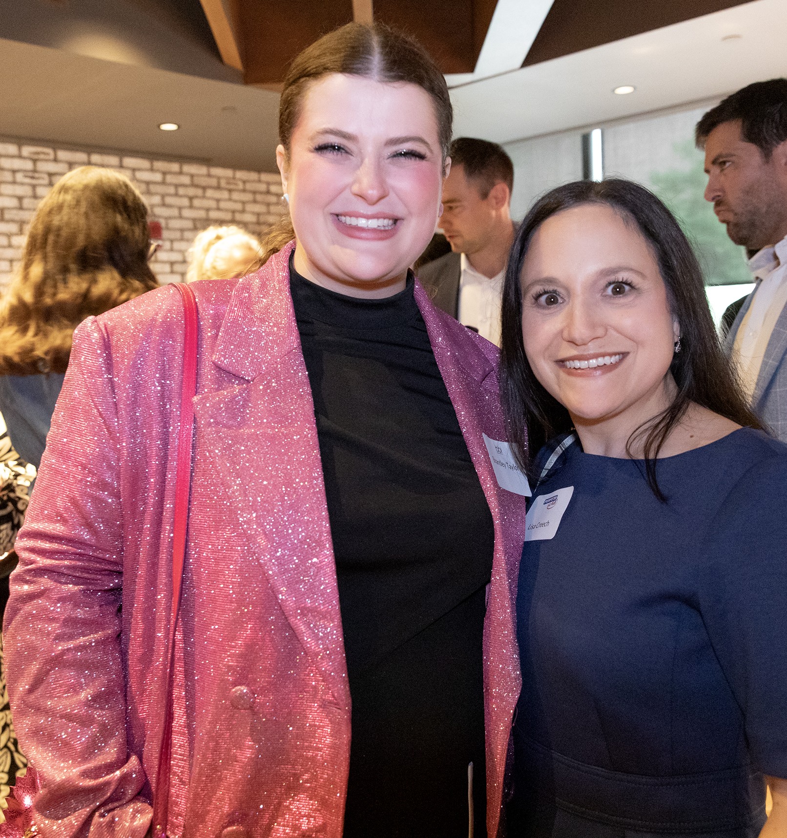 Two women with name tags smile close together at a reception, one in a pink sparkly blazer.