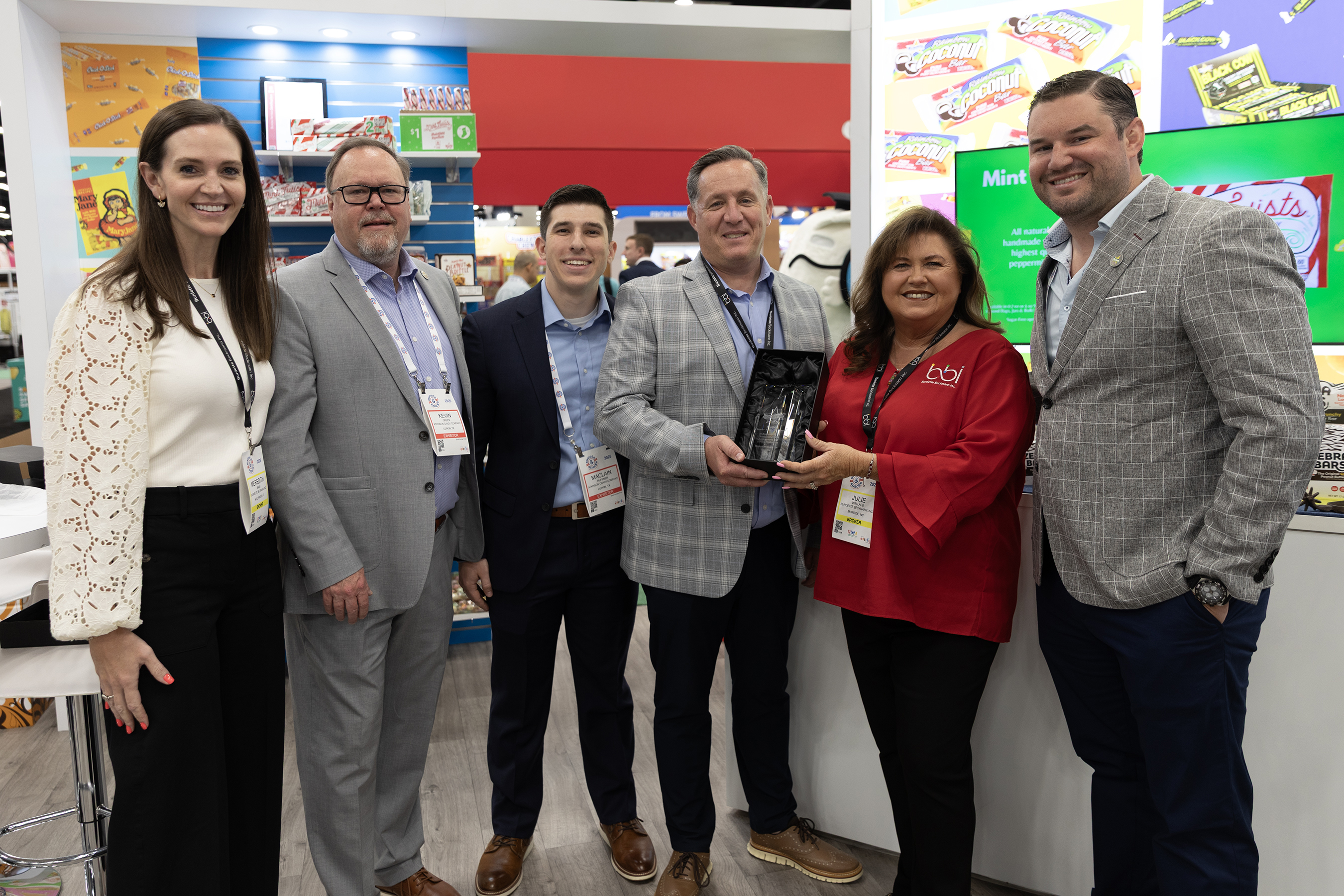 Six adults pose at a candy booth, one man holding a glass award beside a woman in a red top with a bbi logo.