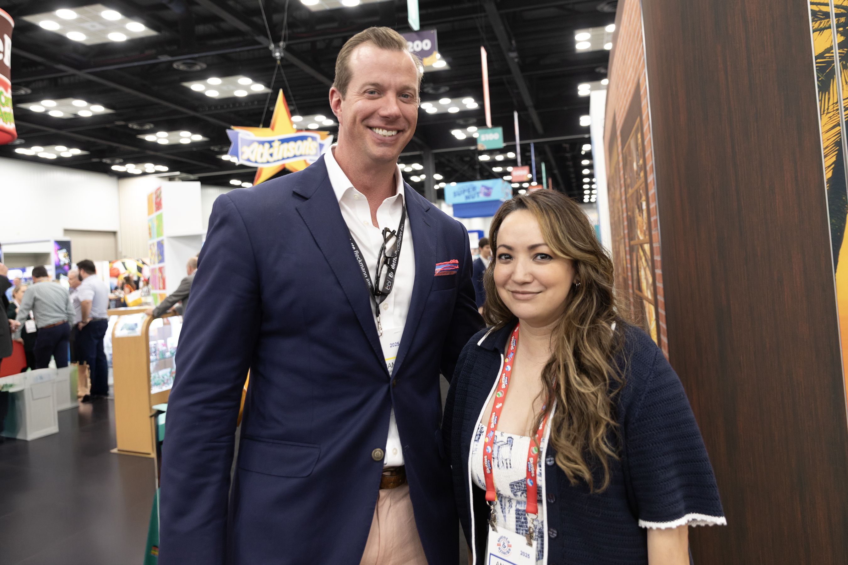 Man in a navy blazer and a woman wearing a lanyard pose together in an expo hall aisle.