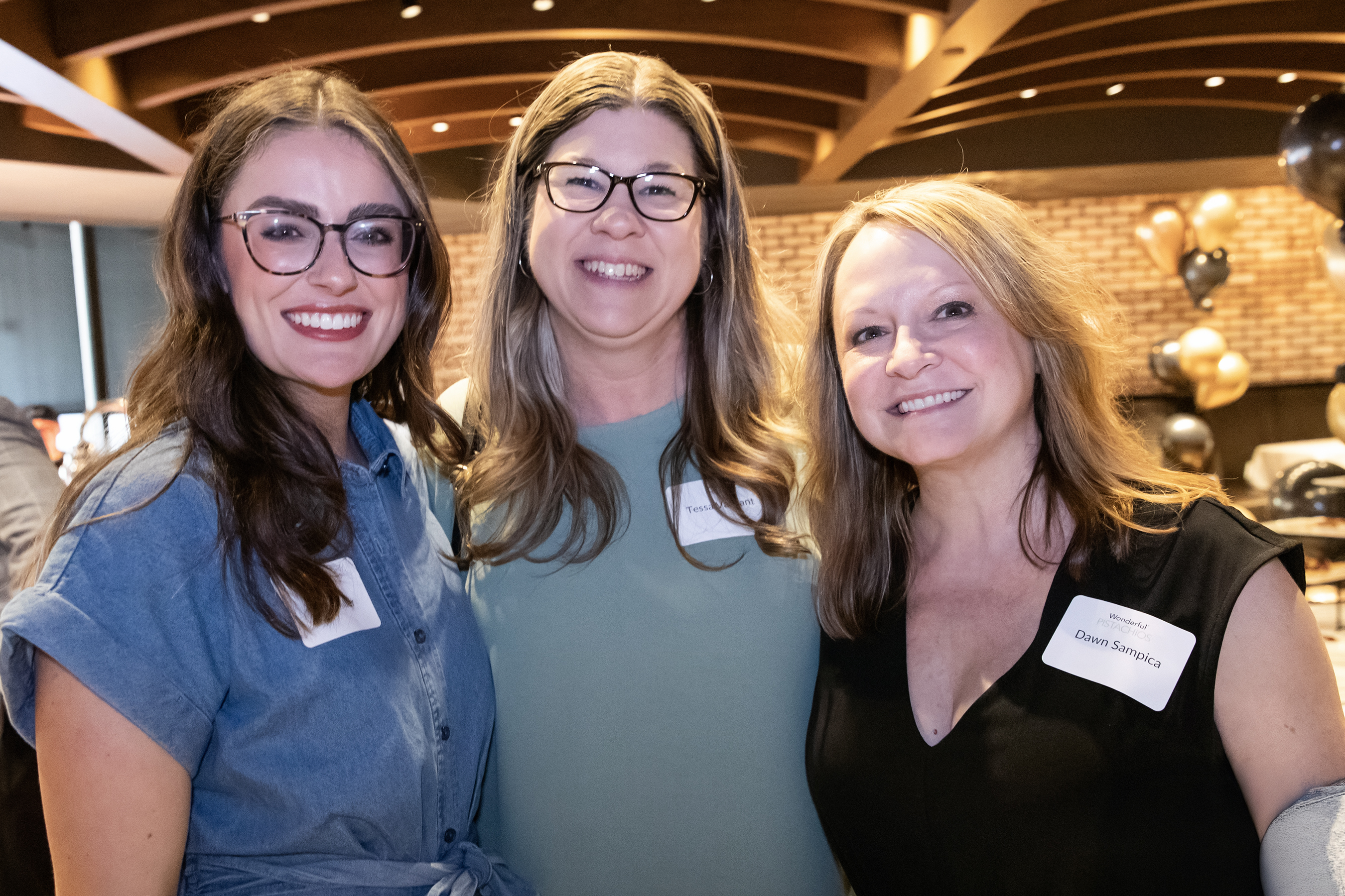 Three smiling women with name tags pose together at a reception, with black and gold balloons behind them.