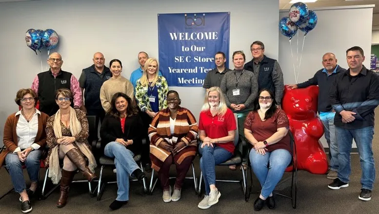 Group of about sixteen people, seated and standing, posing in front of the Yearend Team Meeting banner by a red gummy bear.