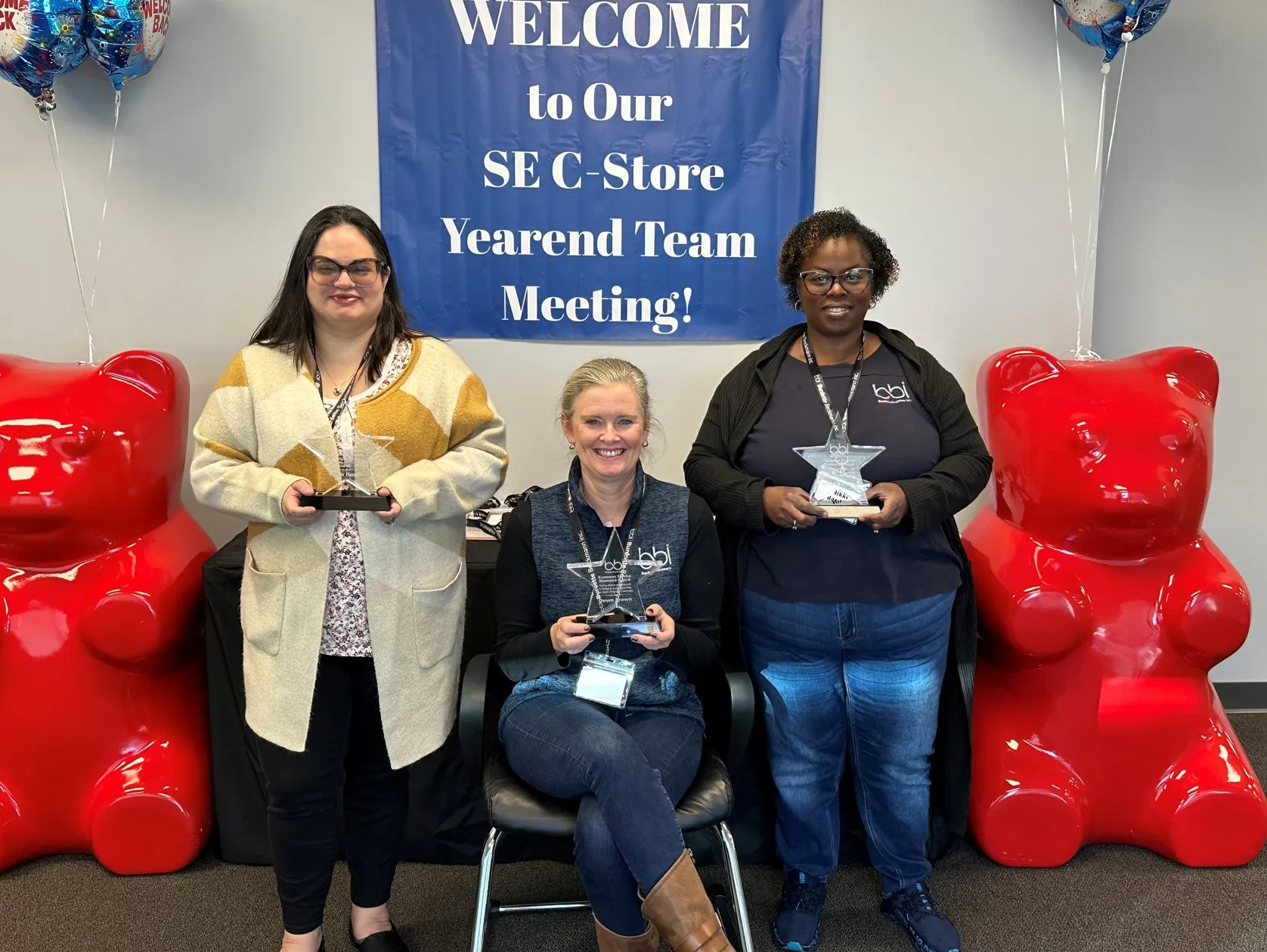 Three women hold glass star awards, one seated, beneath the Yearend Team Meeting banner between red gummy bear statues.