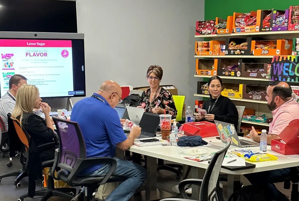 People at a table with laptops and candy boxes, with a screen reading Lower Sugar All The Flavor beside a candy shelf.