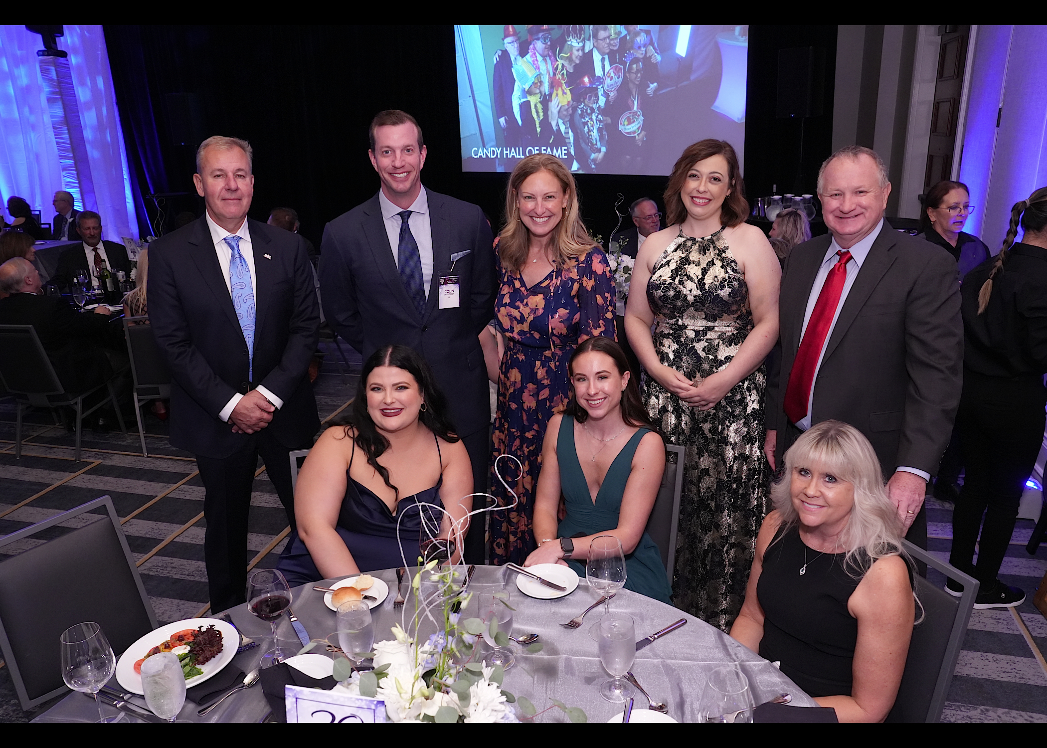 Eight guests in formal wear pose around a banquet table in a ballroom, a Candy Hall of Fame screen behind them.