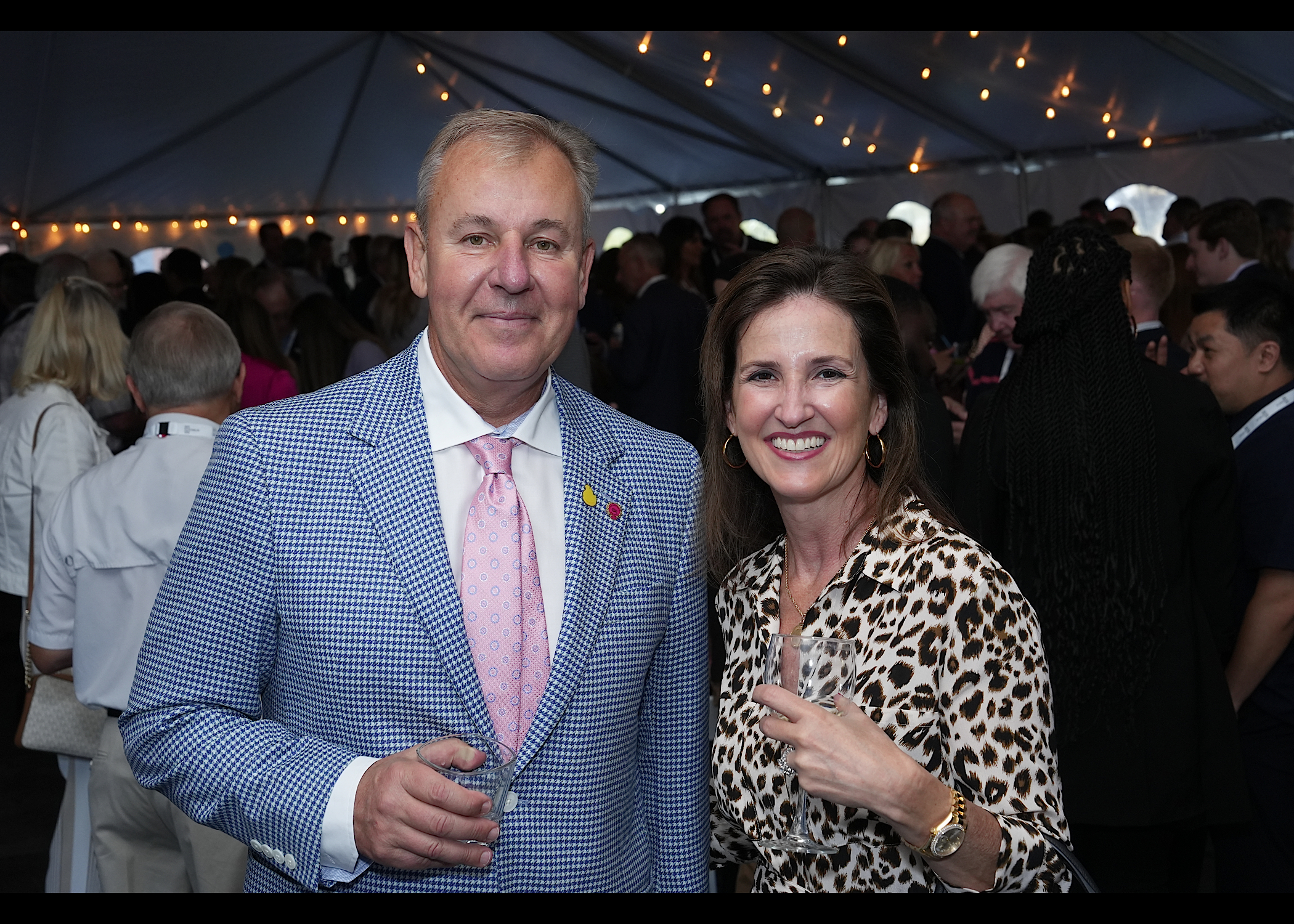Man in a blue houndstooth blazer and pink tie with a woman in a leopard-print top, holding drinks in a tent.
