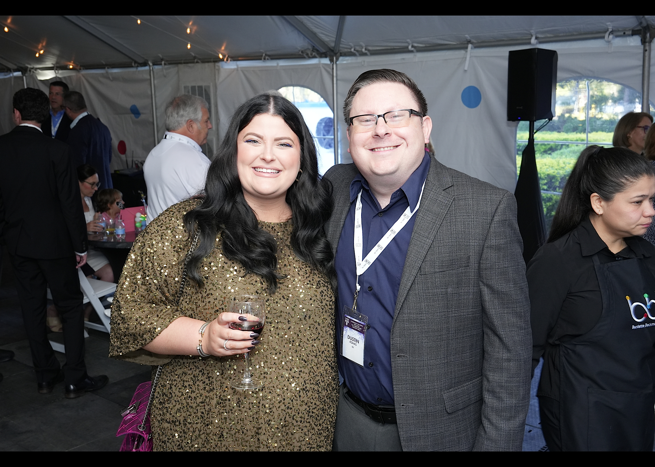 Woman in a gold sequin top and man with glasses in a gray blazer pose in a tent, a server in a bbi apron behind them.
