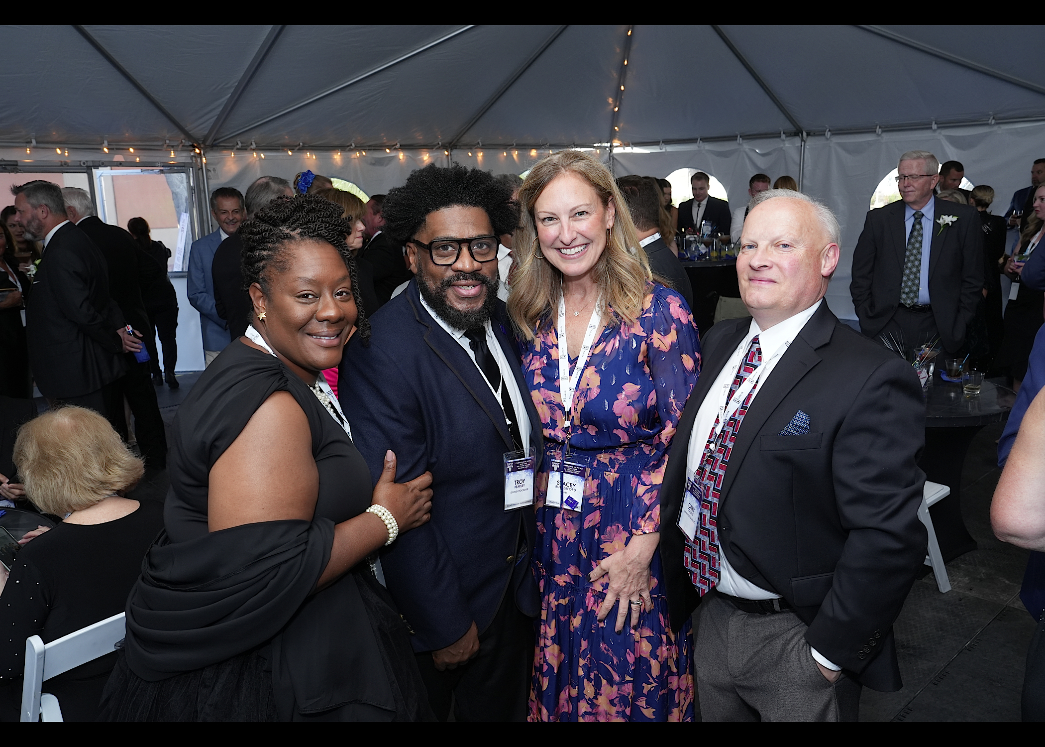 Four guests pose in a tent: a woman in black, a man with glasses, a woman in a floral dress and a man in a red tie.