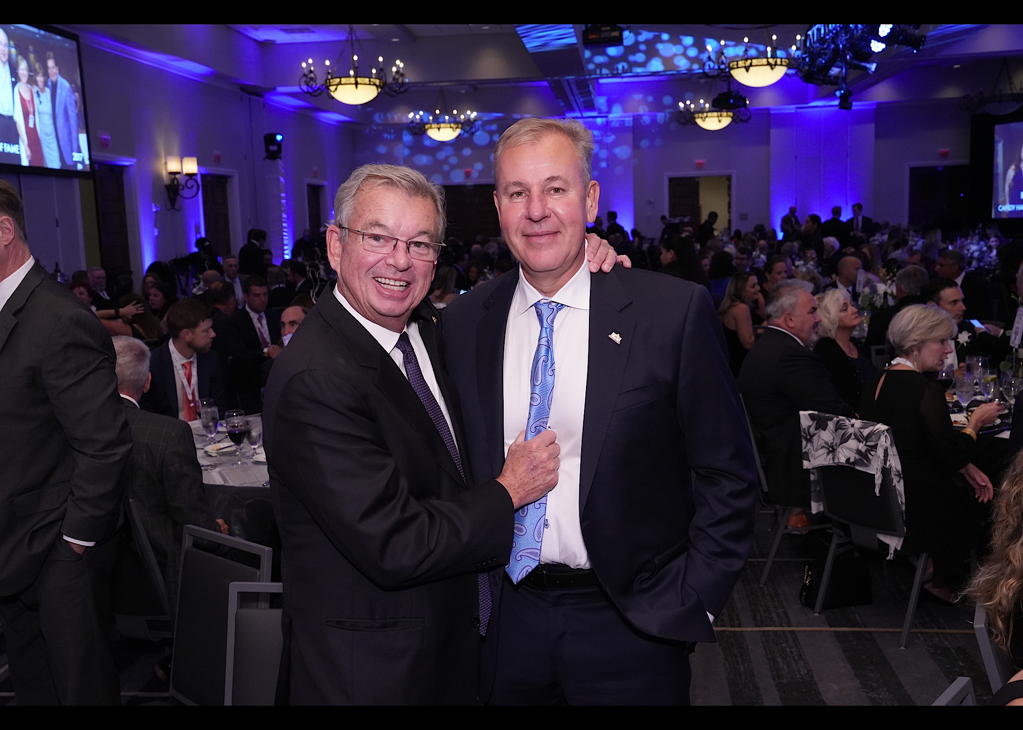 Two men in dark suits, one with an arm around the other, in a blue-lit banquet hall full of seated guests.