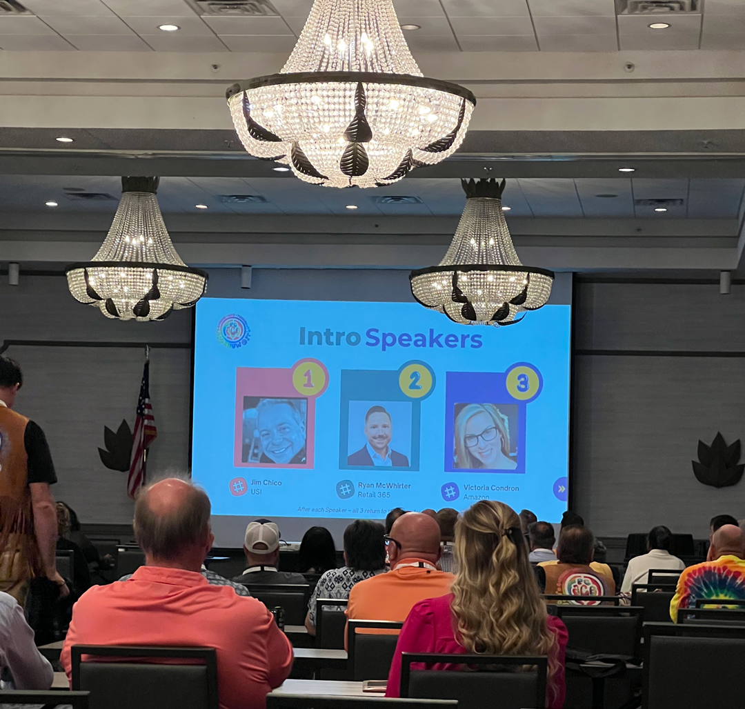 Audience in a ballroom with chandeliers faces a screen titled Intro Speakers showing three speaker portraits.