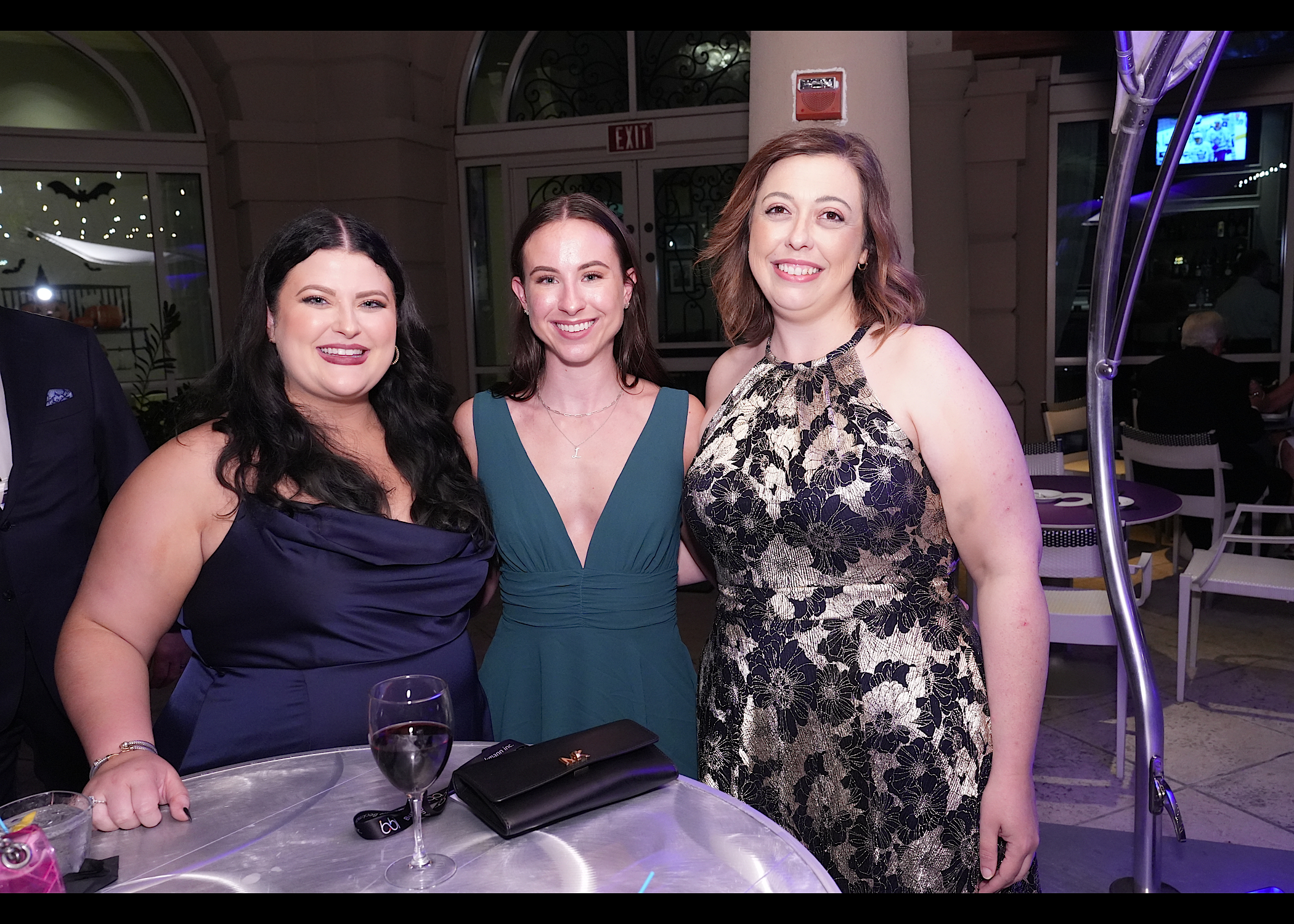 Three women in evening dresses pose at a cocktail table beside a patio doorway.