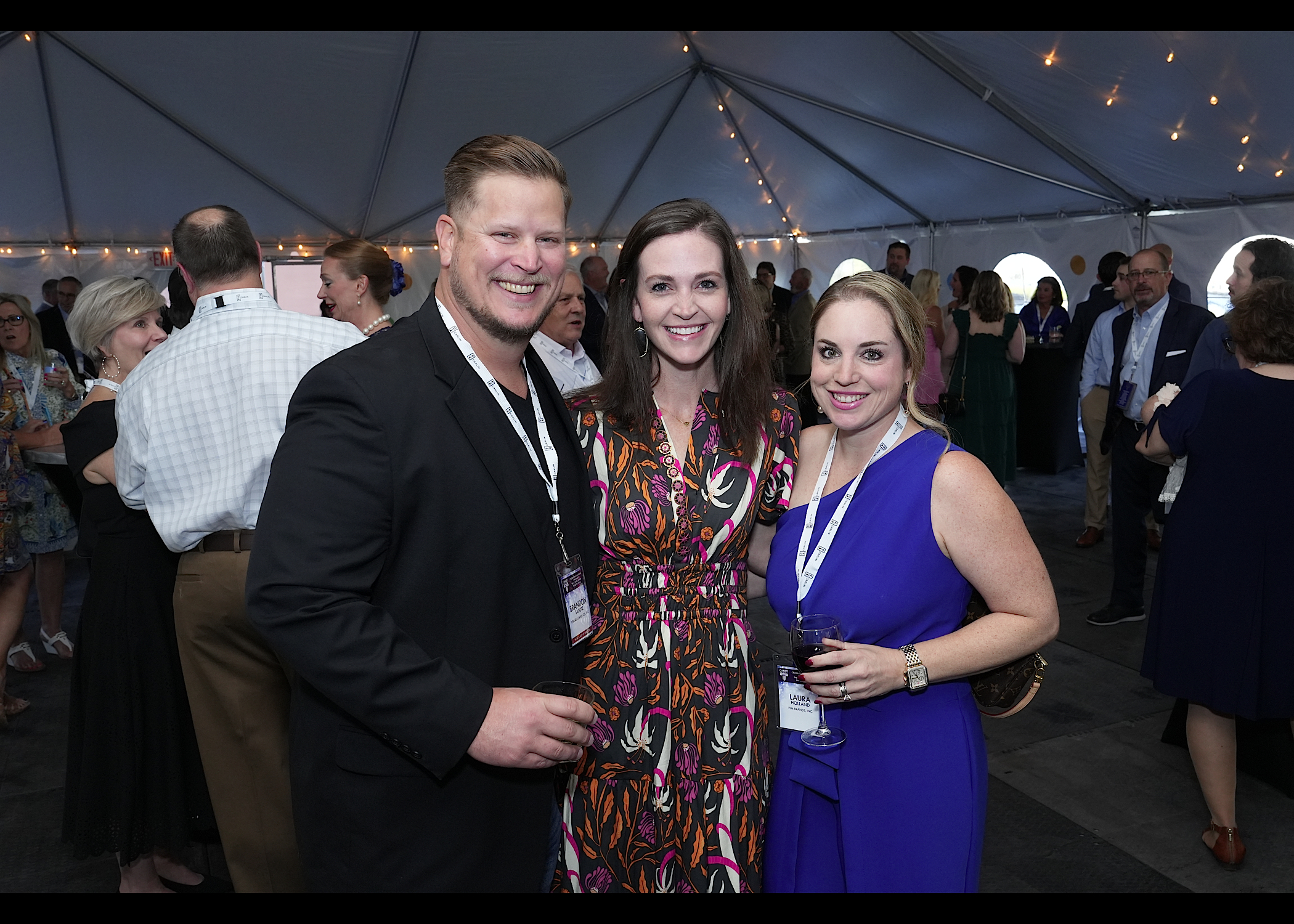 Man in a black blazer with two women in a print dress and a blue dress smiling in a lit tent, holding drinks.