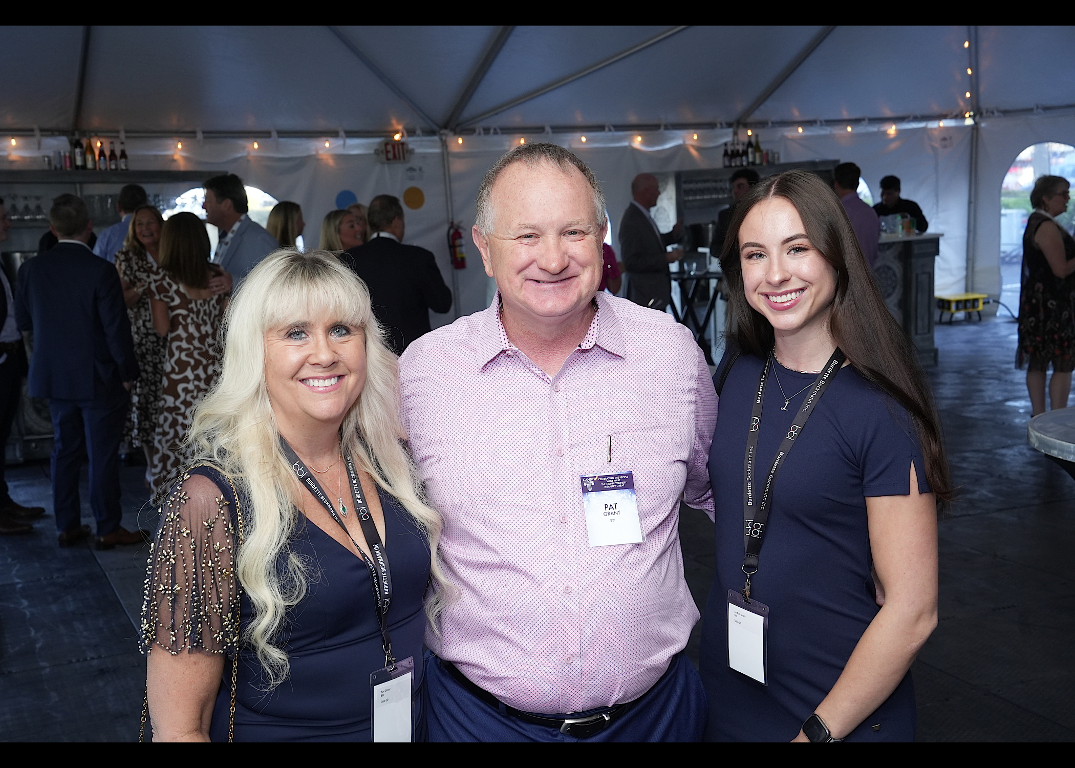 Man in a pink shirt with a name badge poses between two women wearing lanyards in a tent.