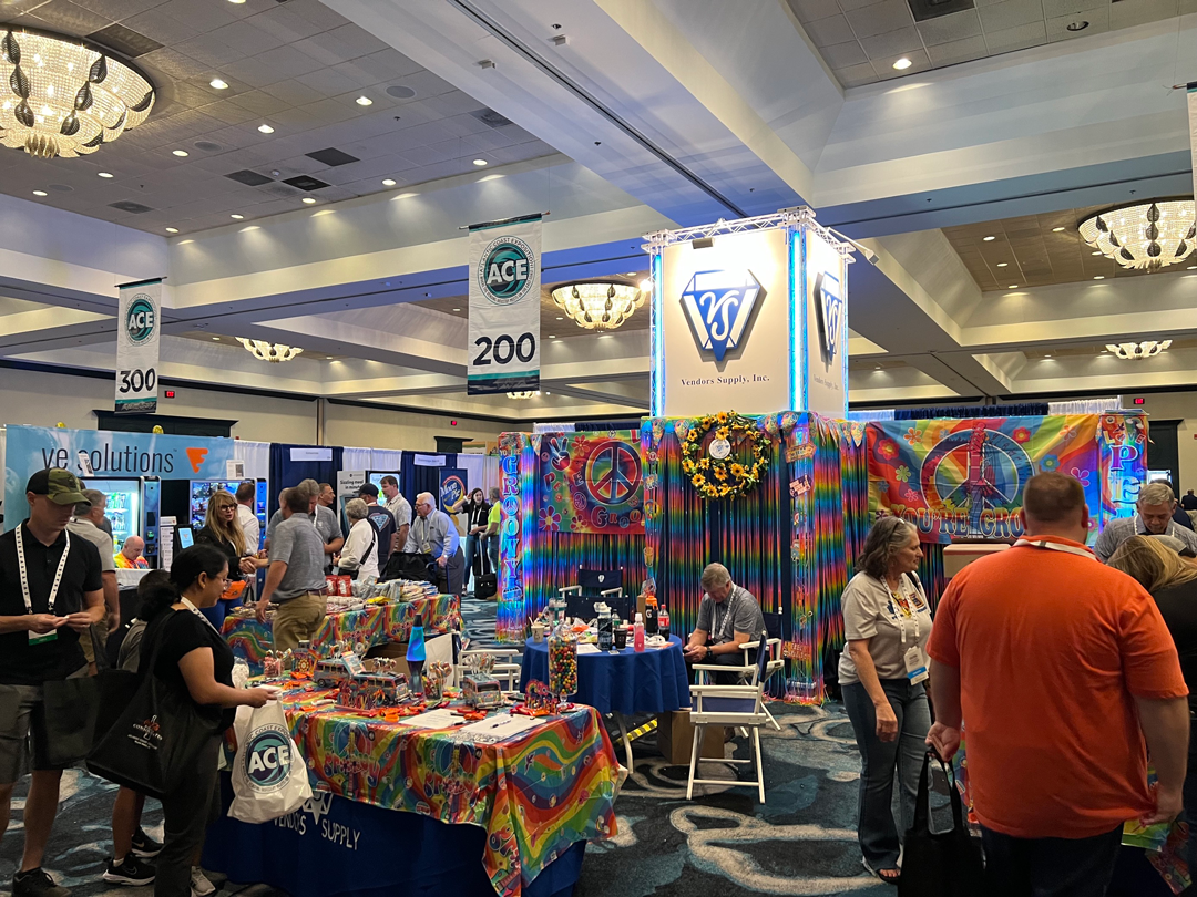 Trade show floor with a tie-dye, peace-sign themed Vendors Supply booth under ACE banners as attendees browse.