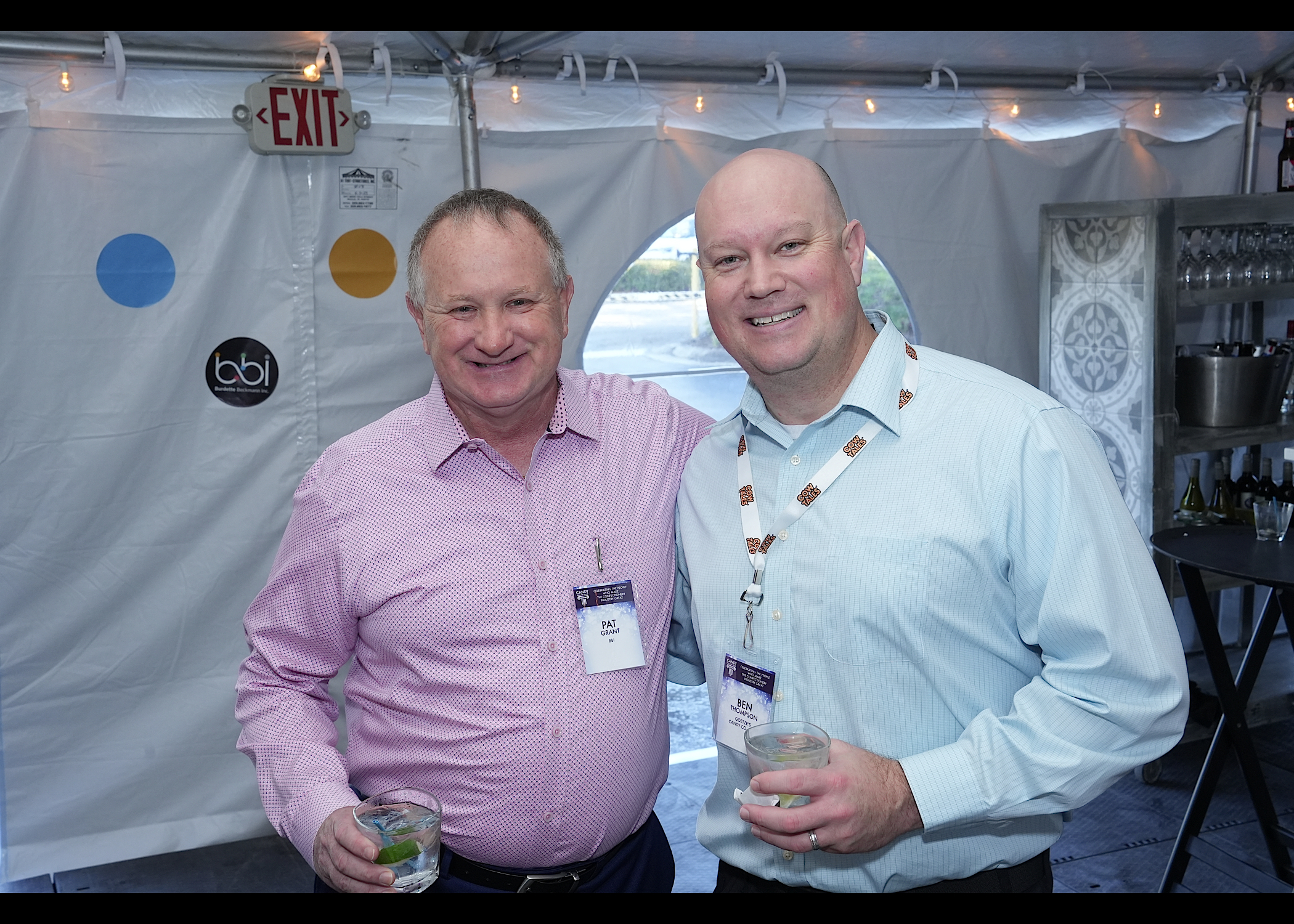 Two men with name badges and drinks pose in front of a tent wall bearing a bbi logo.