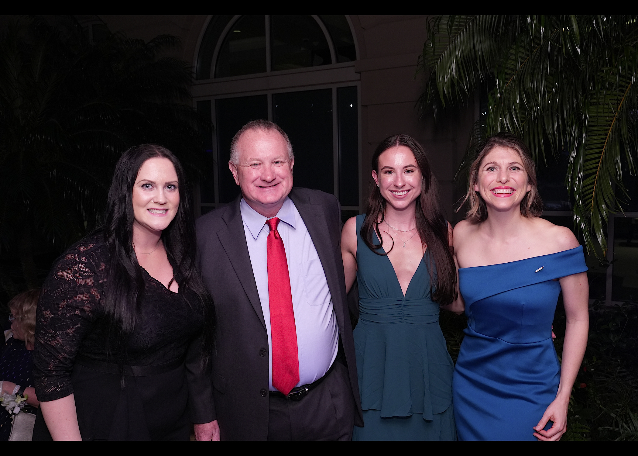 Man in a suit with a red tie and three women in evening dresses pose together at night beside palm leaves.