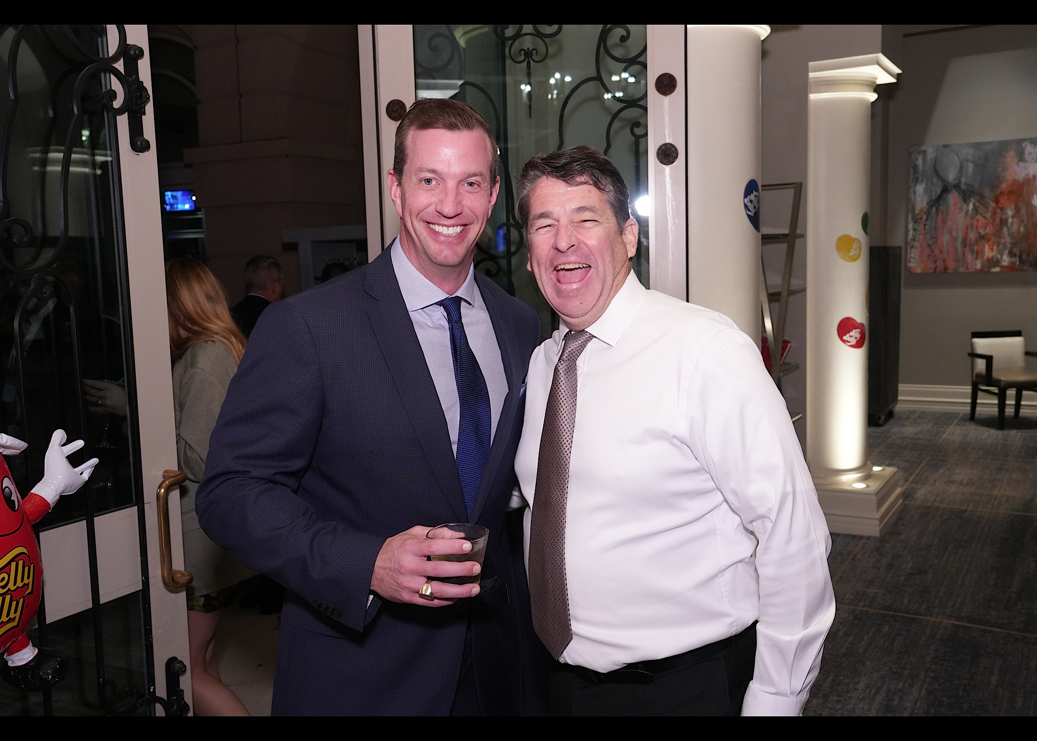 Two men laughing together in a hallway beside a Jelly Belly bean mascot figure.