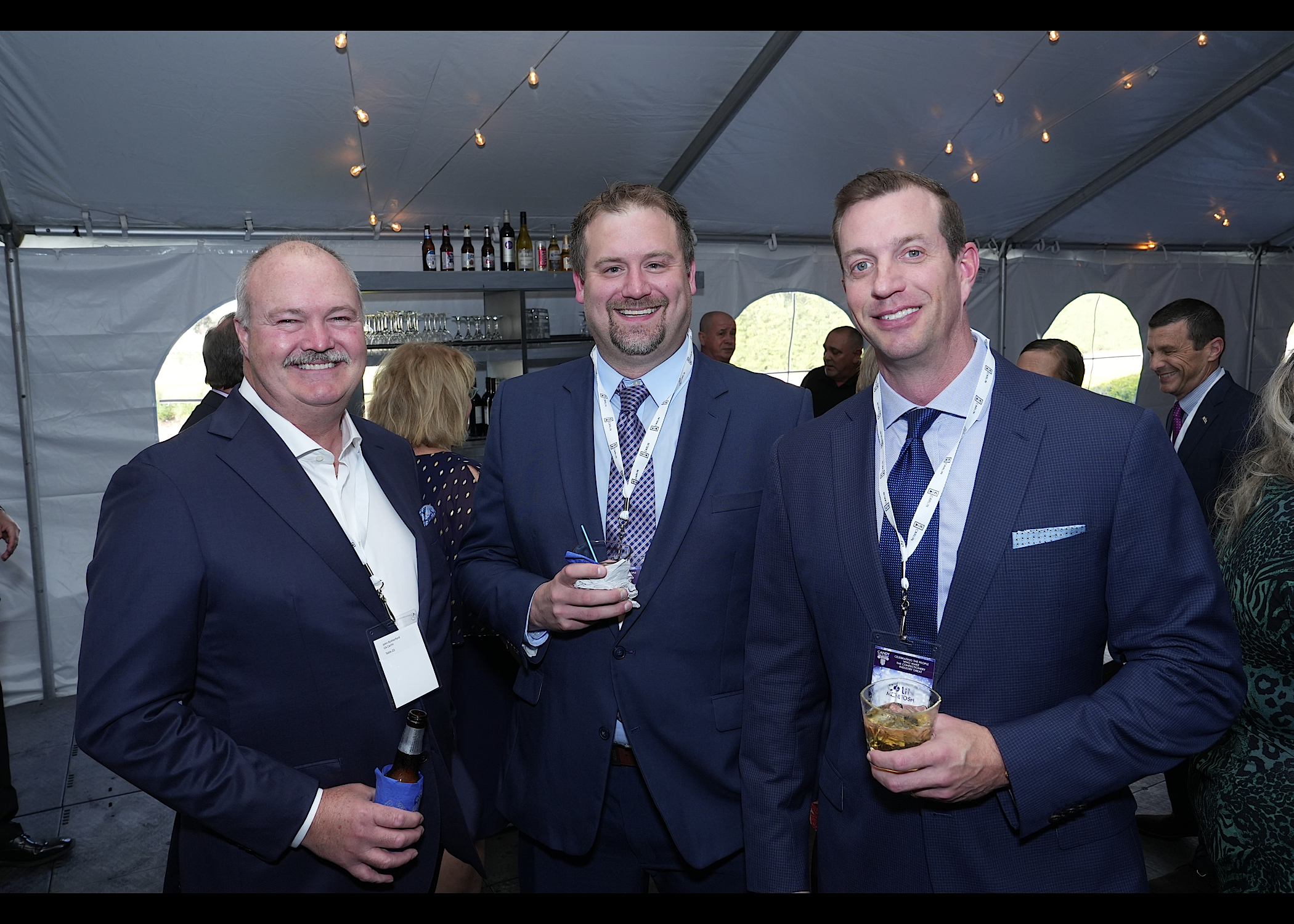 Three men in navy suits and lanyards pose together in a tent, one holding a beer bottle and one a small cup.