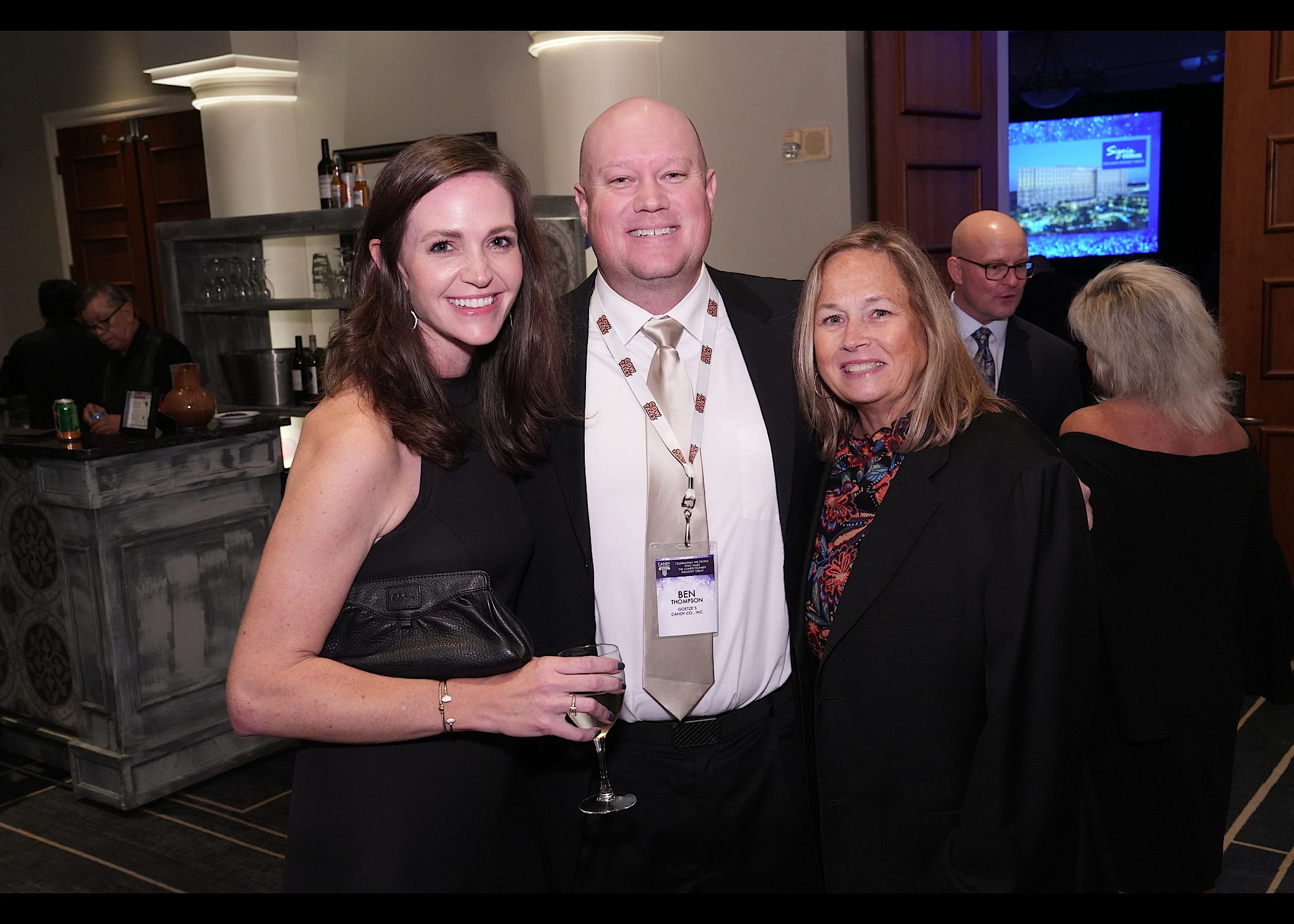 Woman in a black dress, a man with a name badge and a woman in a dark jacket pose in front of a bar.