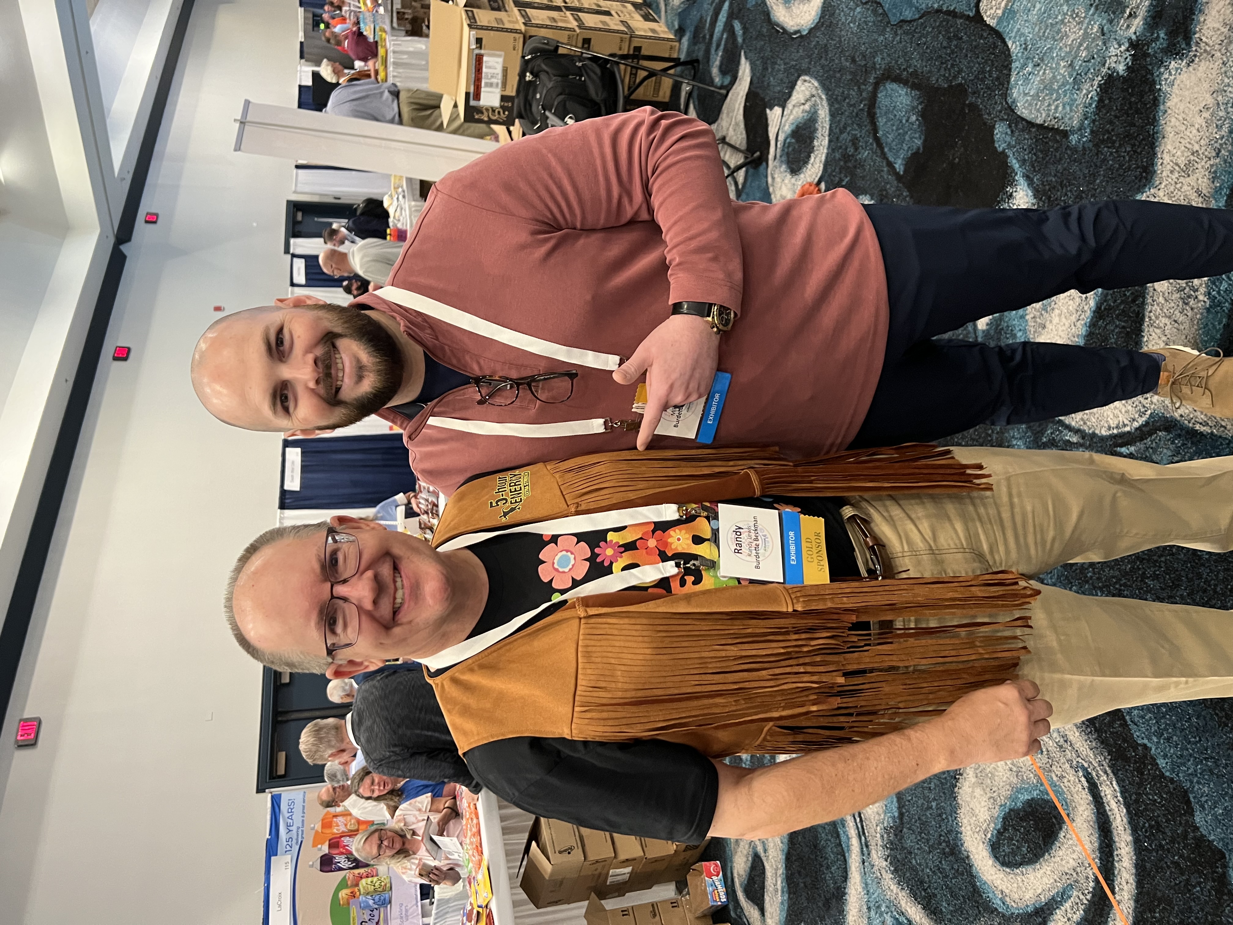 Two men with exhibitor badges pose in a convention aisle, one in a fringed leather vest with floral shirt, shown sideways.