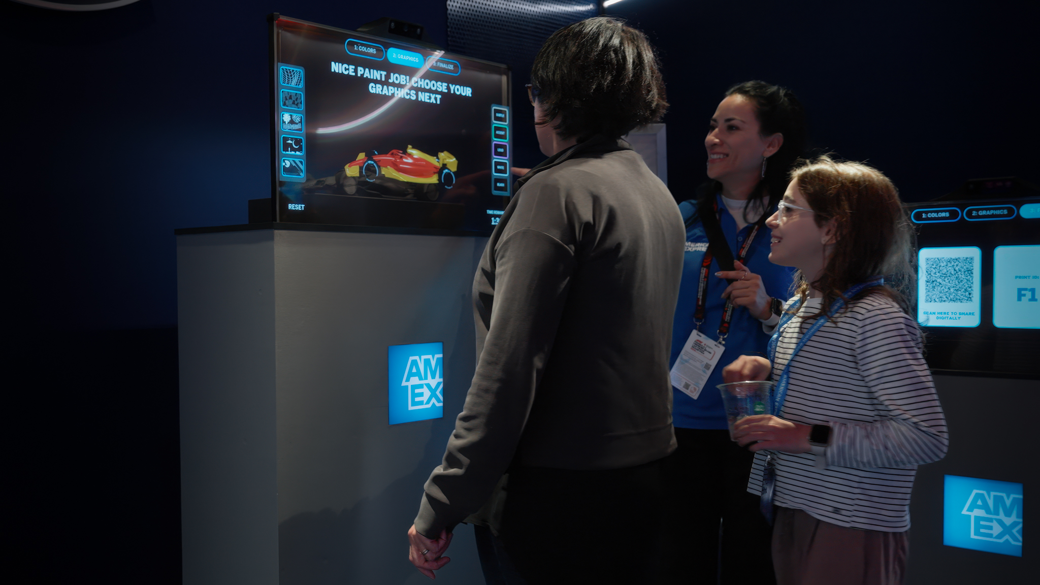 A group of people interacting with a touchscreen kiosk to customize a Formula 1 car livery at the American Express Livery Designer exhibit.