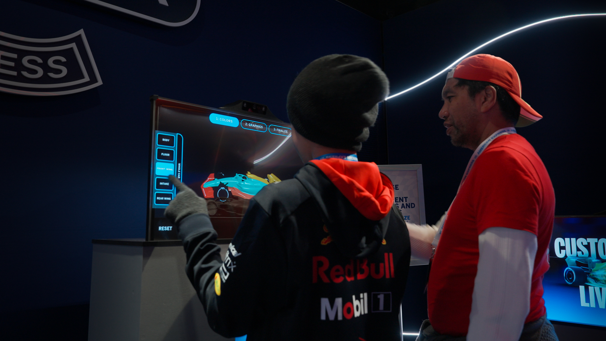 "Two people interacting with a touchscreen display to customize an F1 car livery at an event, with a Red Bull Racing jacket visible."