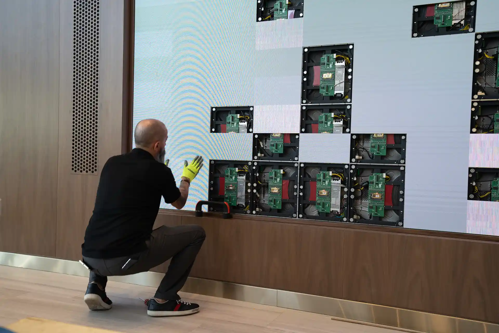 Person installing electronic components on a large display wall, using tools while crouching.