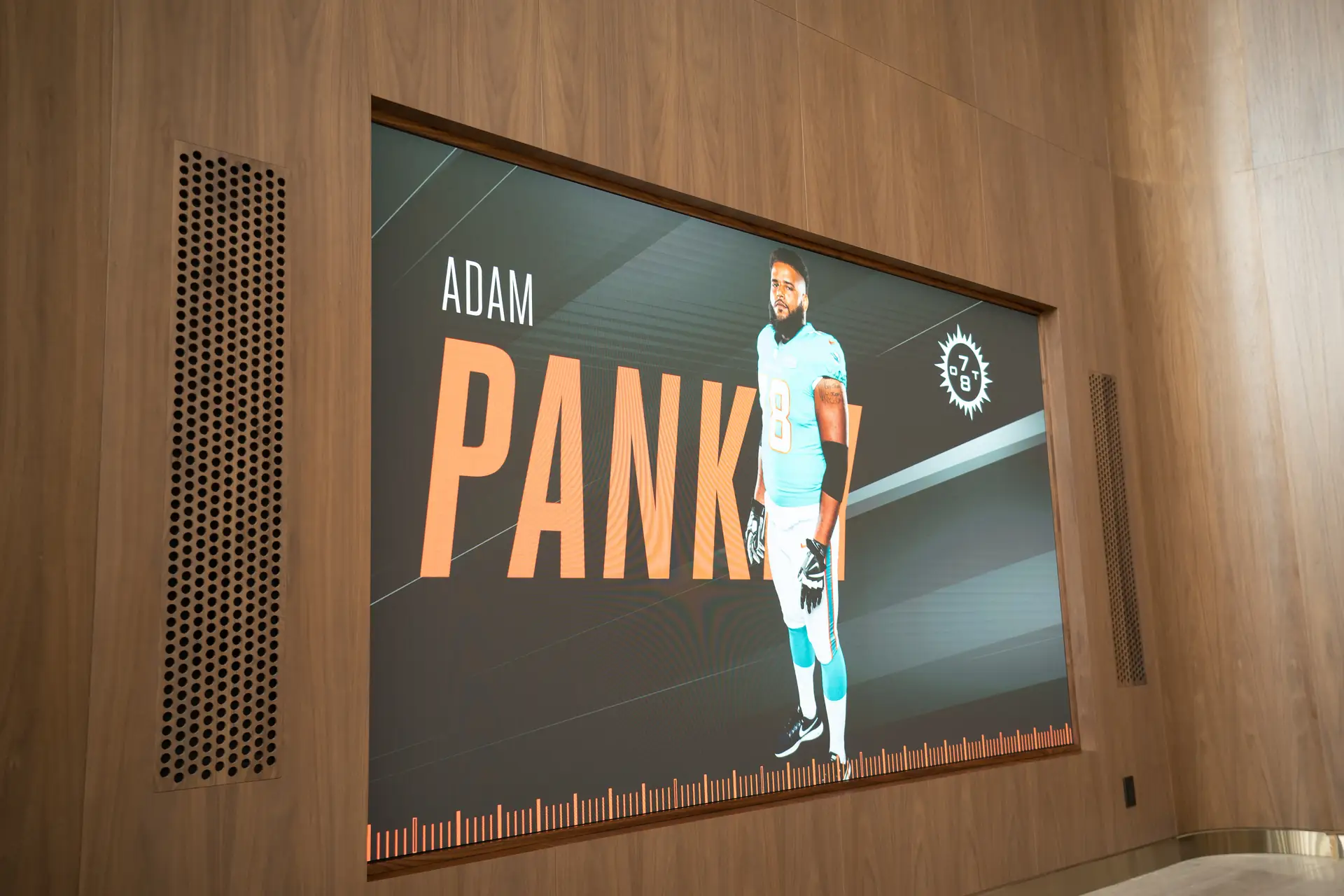 Digital display featuring Miami Dolphins player Adam Pankey, with bold orange text on a wooden wall inside a modern space.