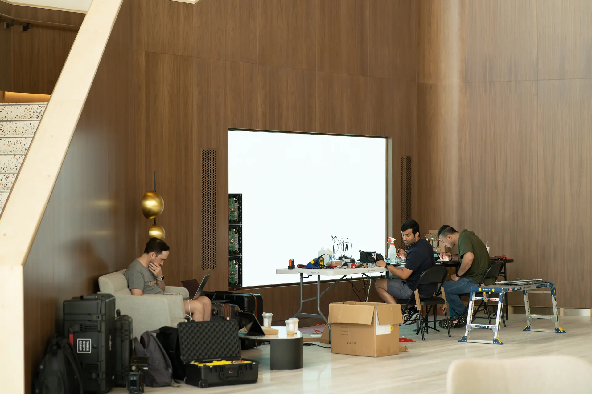 People setting up equipment in a modern room with a wooden wall, a staircase, and a large white screen display.