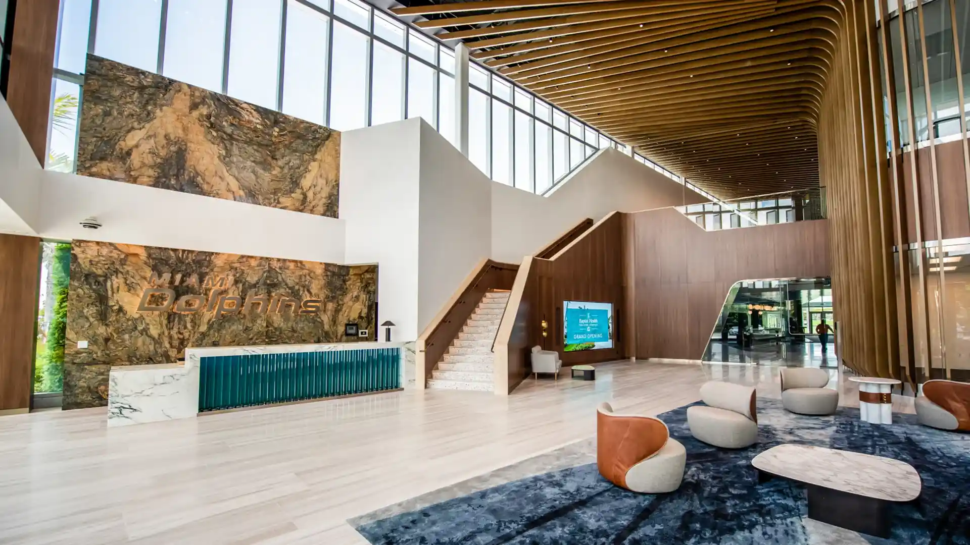 Modern Miami Dolphins reception area with a marble backdrop, teal desk, wooden staircase, and digital display of player Malcolm Perry.