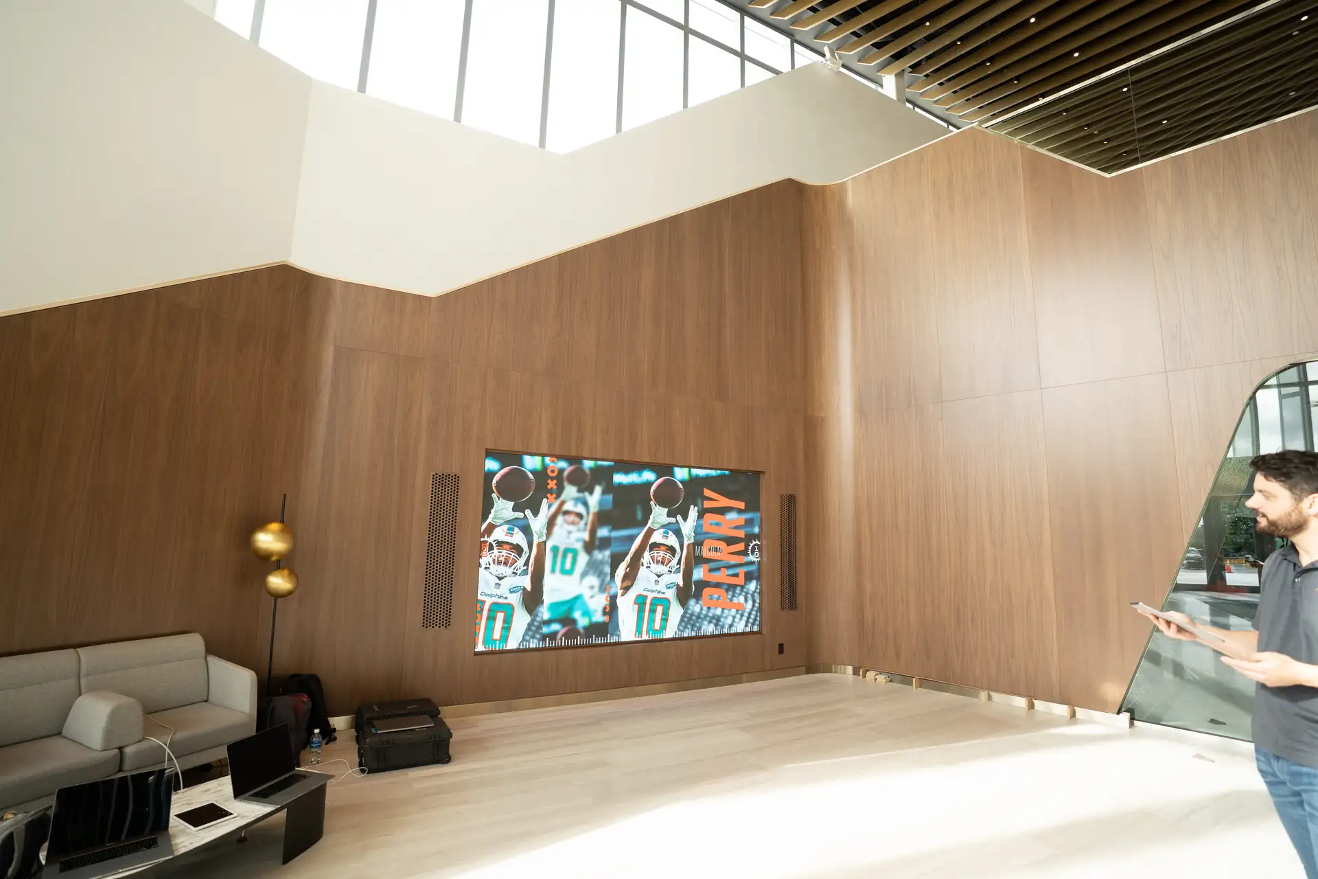 Spacious room with a wooden wall, large digital display, and seating area, illuminated by natural light from large windows above.