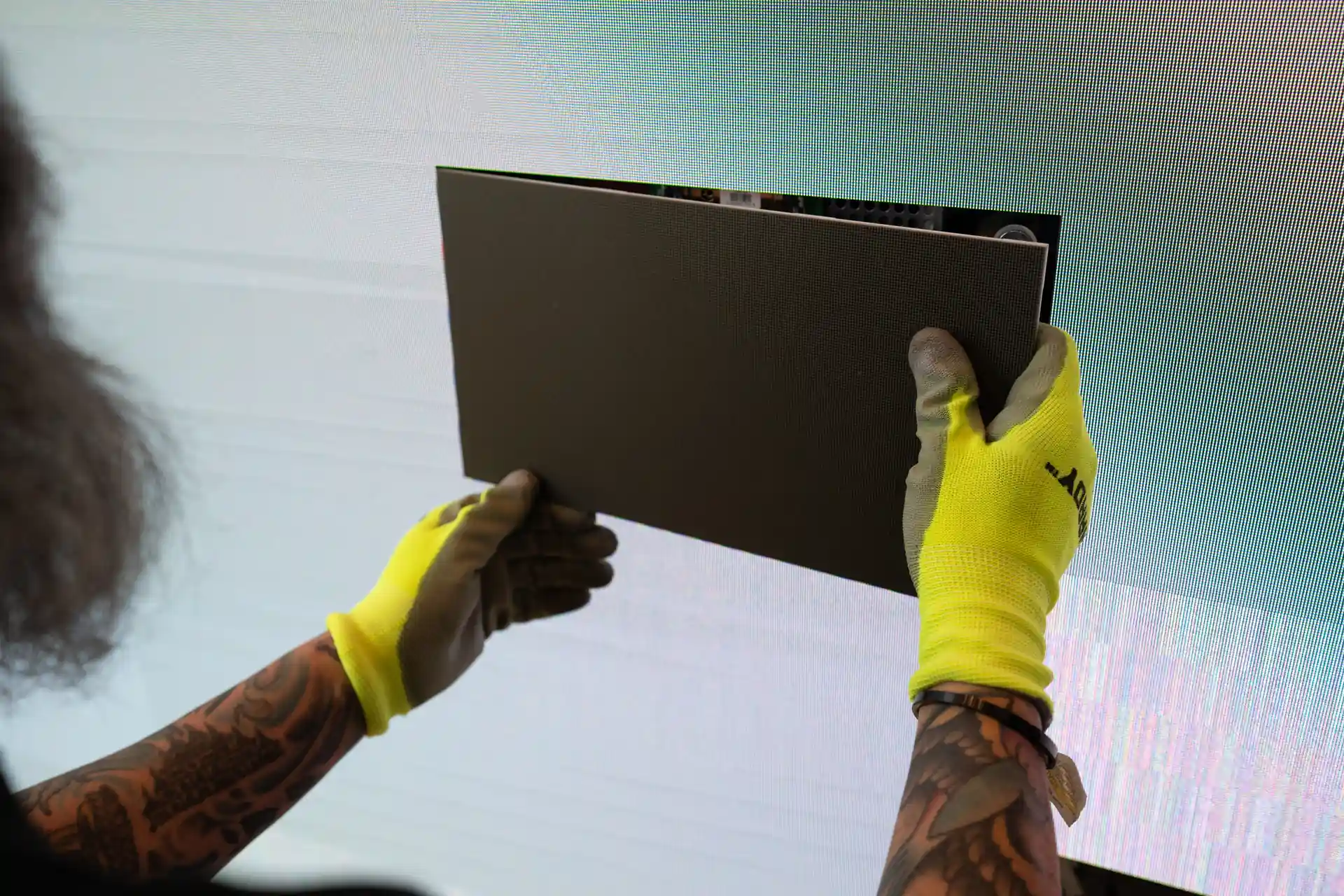 Person wearing yellow gloves holding a rectangular led panel up to an empty spot in the screen