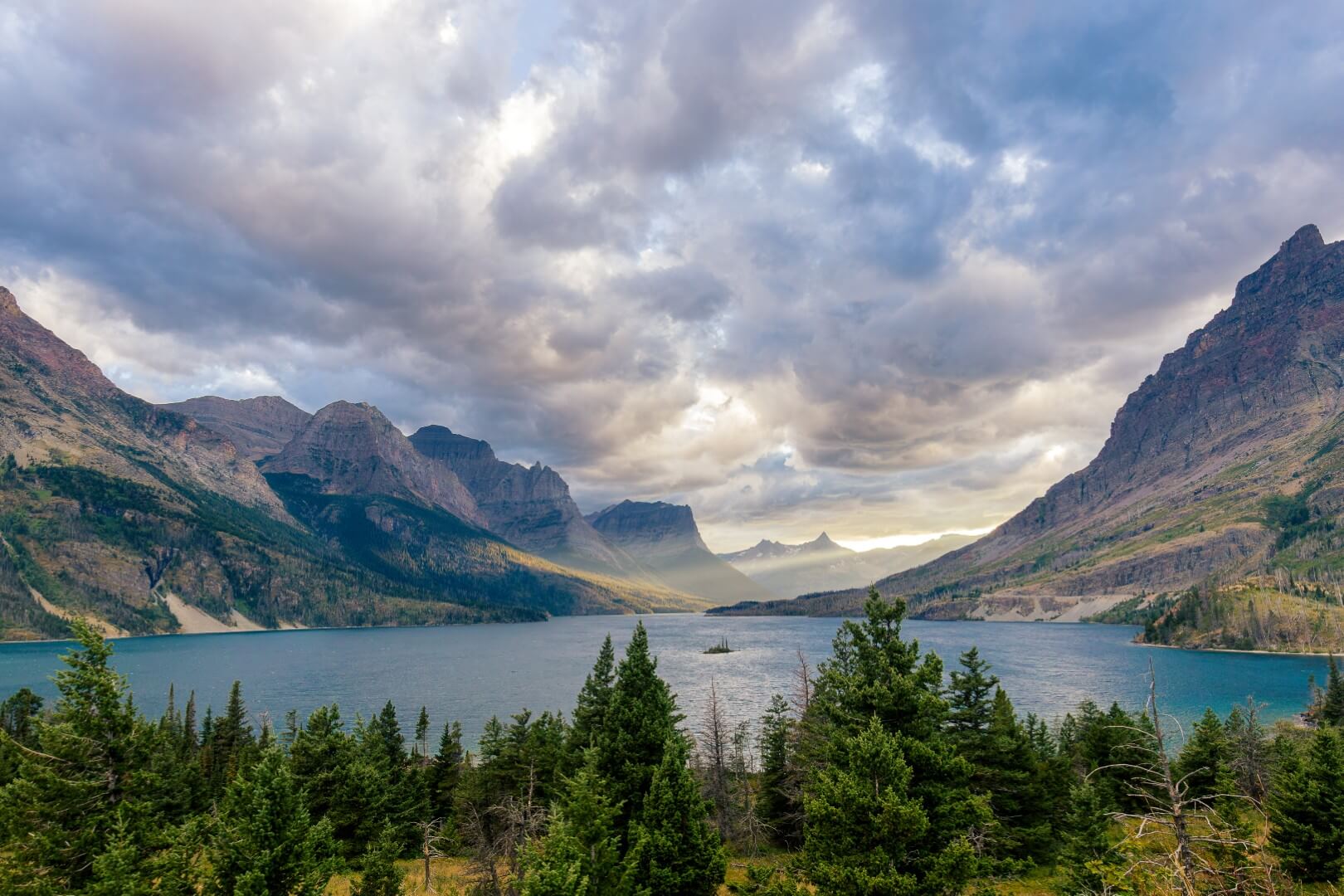 A dramatic sunset in Glacier National Park looking out at a small island in a lake with colorful clouds above and a beam of sunlight breaking through
