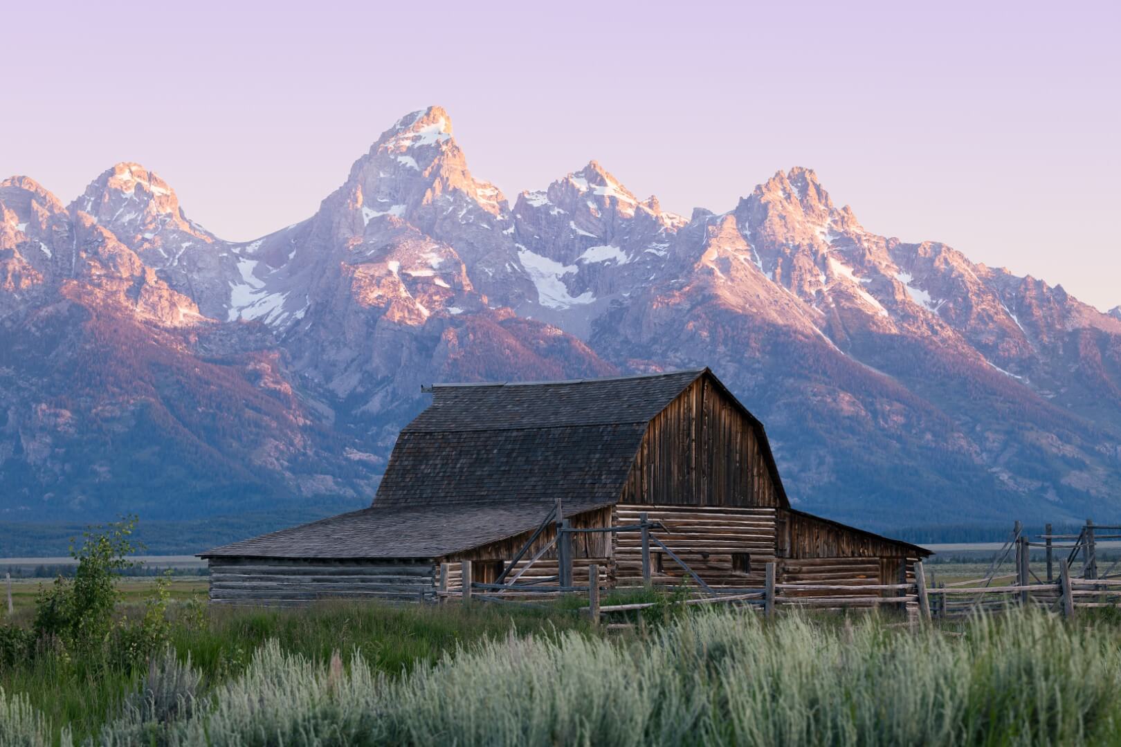 A sunrise photo of an historic barn in Grand Teton National Park with the mountain range light up colorfully in the distance