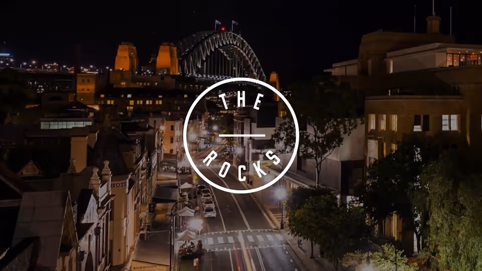 Night view of a city street with illuminated buildings and the Sydney Harbour Bridge in the background, with a white circular logo reading 'THE ROCKS' overlaying the scene.