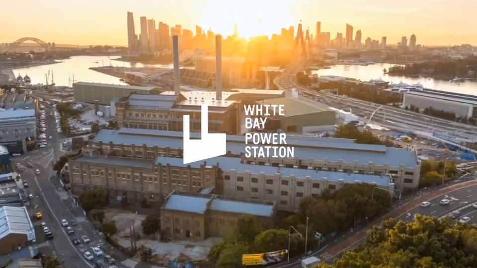 Aerial view of White Bay Power Station building at sunset with city skyline and Sydney Harbour Bridge in the background.