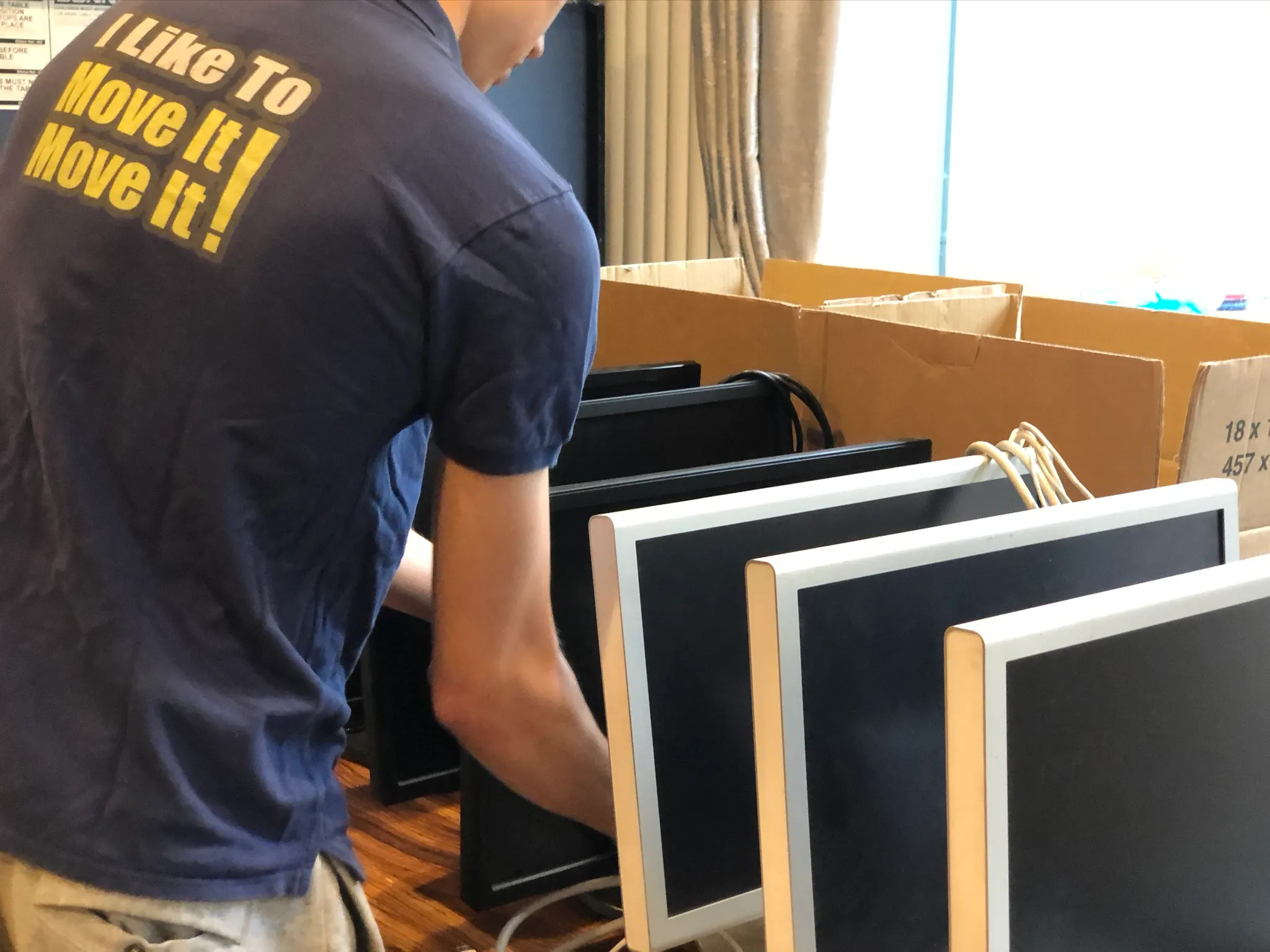 Person in a blue shirt that says 'I Like To Move It! Move It!' arranging multiple black computer monitors with white frames inside large cardboard boxes.