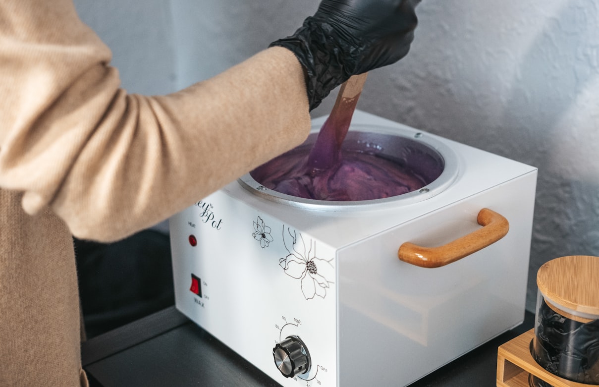 Gloved esthetician stirring warm hair-removal paste in a pot during a professional treatment